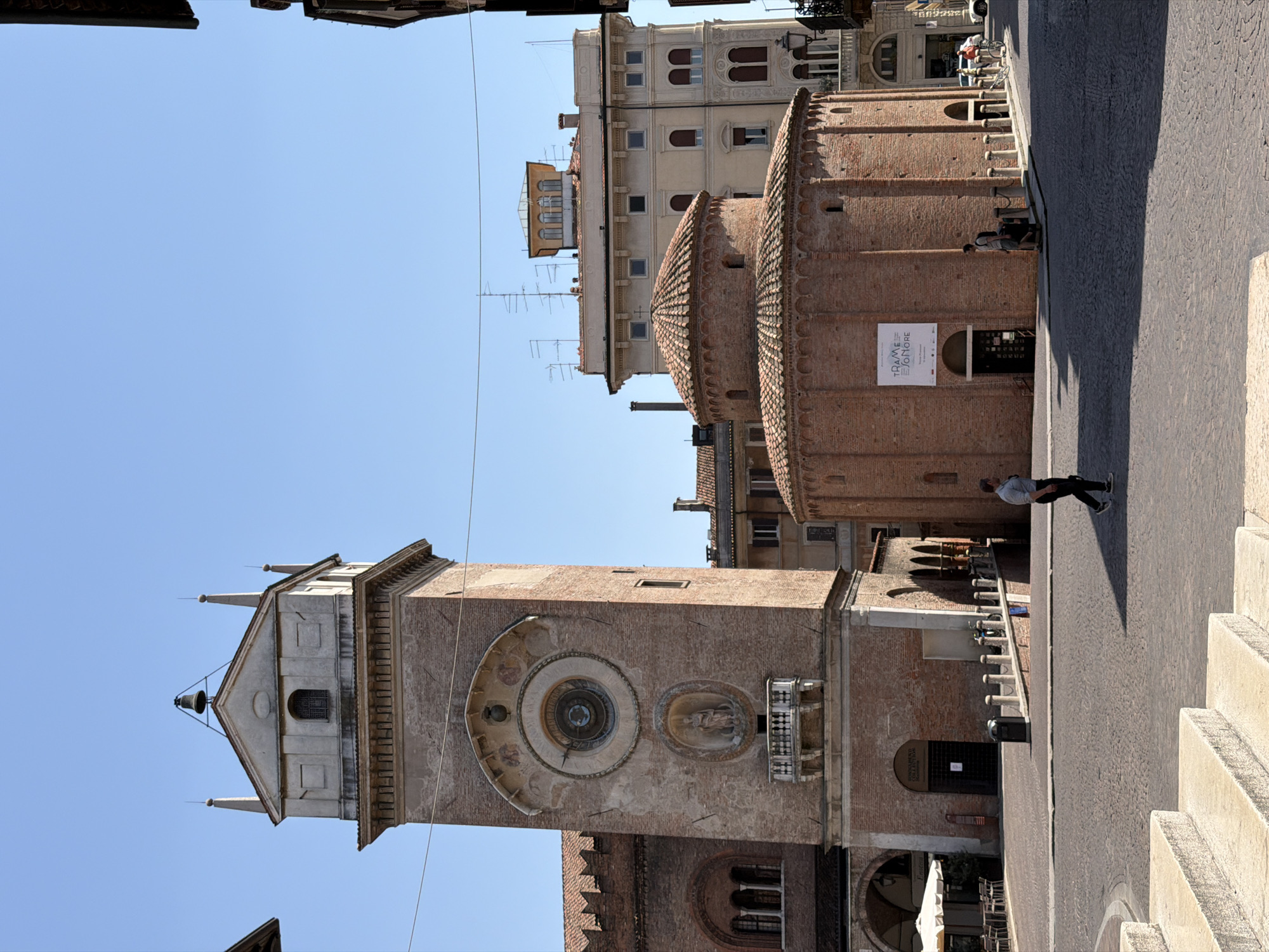 The image shows a street view in Mantua, Italy. The architectural structures present are primarily constructed of brick and stone. The Torre dell'Orologio (Clock Tower) is prominent, with visible details like the astronomical clock face and statues. Adjacent to the tower is the Rotonda di San Lorenzo, a round brick church. The buildings suggest a historic area. A person is walking along the street, which appears to be paved with stones or cobblestones. The sky is clear. There are other buildings in the background.