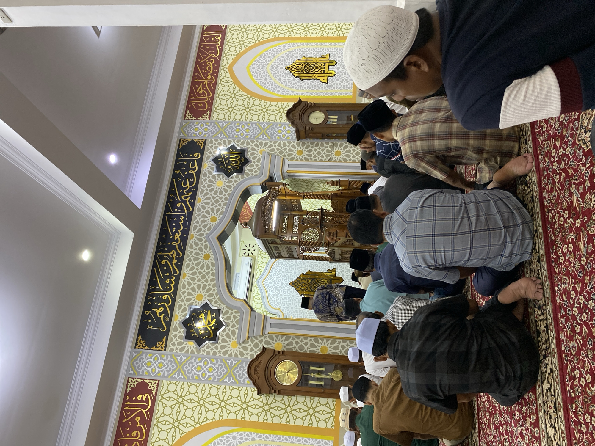In Lhokseumawe, Indonesia, the interior of a mosque is depicted with worshippers seated on a red carpet during prayer. Men, predominantly wearing skullcaps and modest attire, are arranged in rows, facing the front of the mosque. In the background, an ornate wooden pulpit stands beneath an arched entryway decorated with Islamic calligraphy and geometric patterns. A grandfather clock is visible to the left, also adorned with decorative elements. The walls are elaborately patterned in gold, white, and red, featuring Arabic script. The lighting appears to be from overhead fixtures, contributing to a warm, ambient glow.
