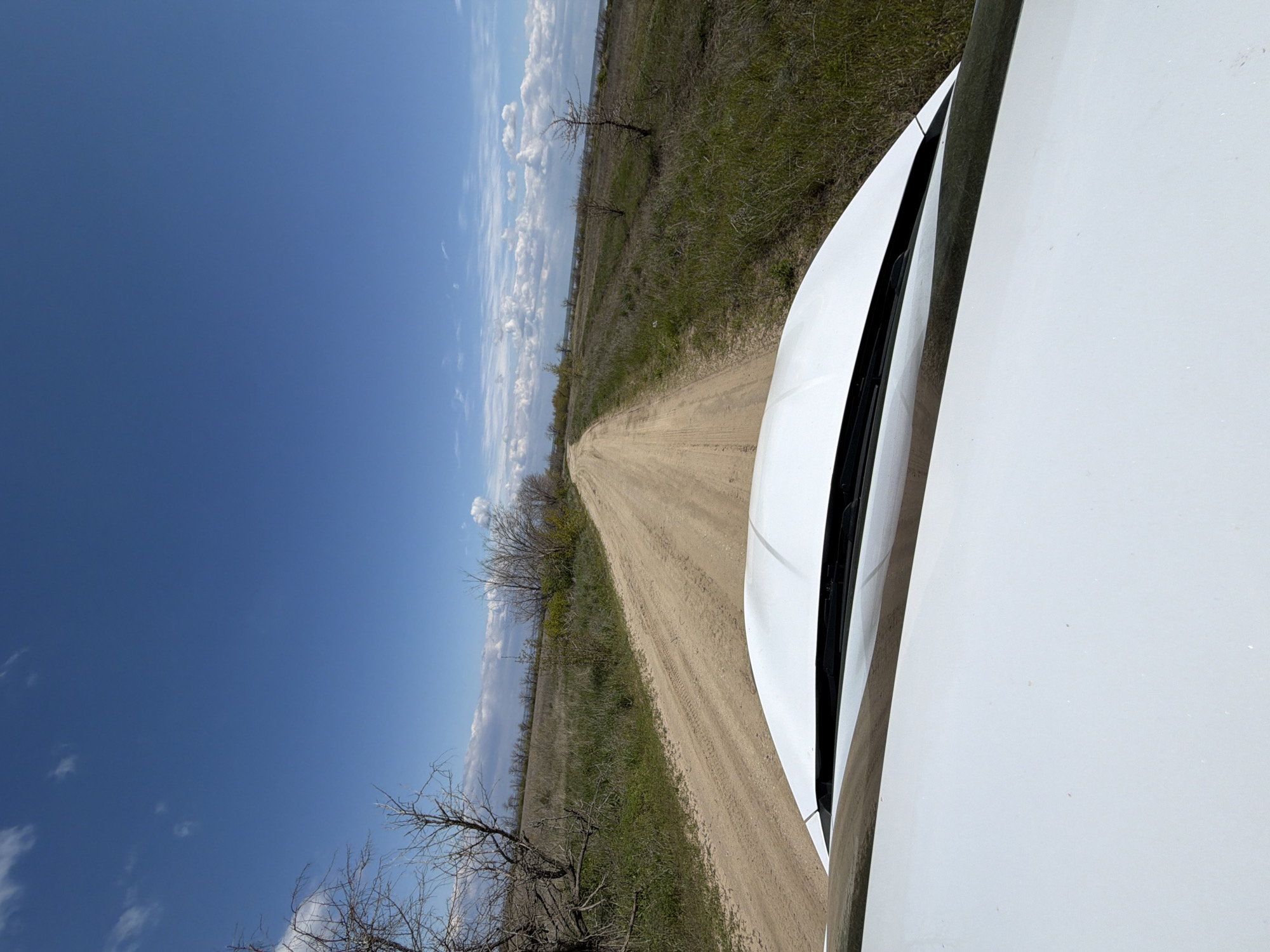 The image shows a dirt road stretching into the distance under a blue sky with scattered clouds. The road is flanked by grassy fields with bare trees. The hood of a white vehicle is visible in the foreground, suggesting the photo was taken from inside the car. The location is likely near Verkh. Lipovka, Russia, based on the prompt.