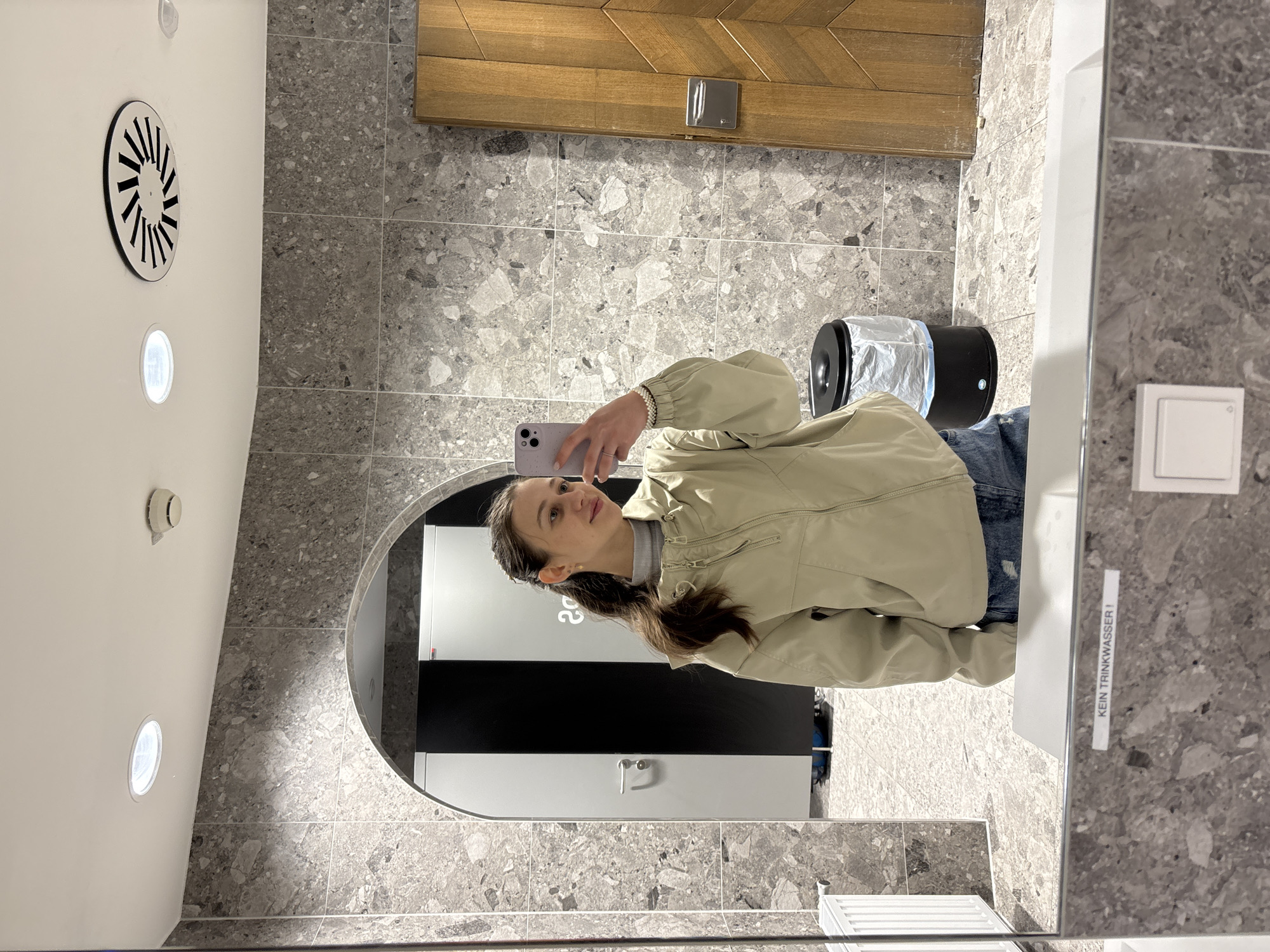A young woman with dark brown hair tied in a ponytail is taking a mirror selfie in a modern restroom. She is wearing a light beige hooded jacket and denim shorts. She is holding a light purple smartphone in her right hand.
The restroom features grey marble-patterned tiles on the walls and floor. To the left of the woman, an arched doorway reveals a glimpse of stalls with black and white doors and a white radiator. A black trash can with a plastic liner is visible to her right. A wooden door with a chevron pattern is also on the right side of the frame. The ceiling has a circular vent and a recessed light fixture. In the reflection of the mirror, a white button and a label that reads "KEIN TRINKWASSER !" are visible. The overall impression is a clean and contemporary public restroom. The location in the city of Ehrwald, Austria is not discernible from the image.