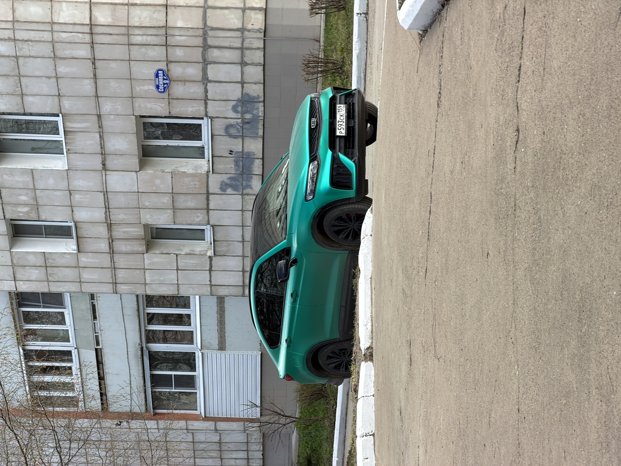 The image shows a green car parked next to a sidewalk and a building with windows and a street sign. The street sign, written in Russian, is “Сосновая 9” (Sosnovaya 9). The car's license plate reads "P593CK159." The location is most likely in the city of Chaikovsky, Russia based on the license plate.