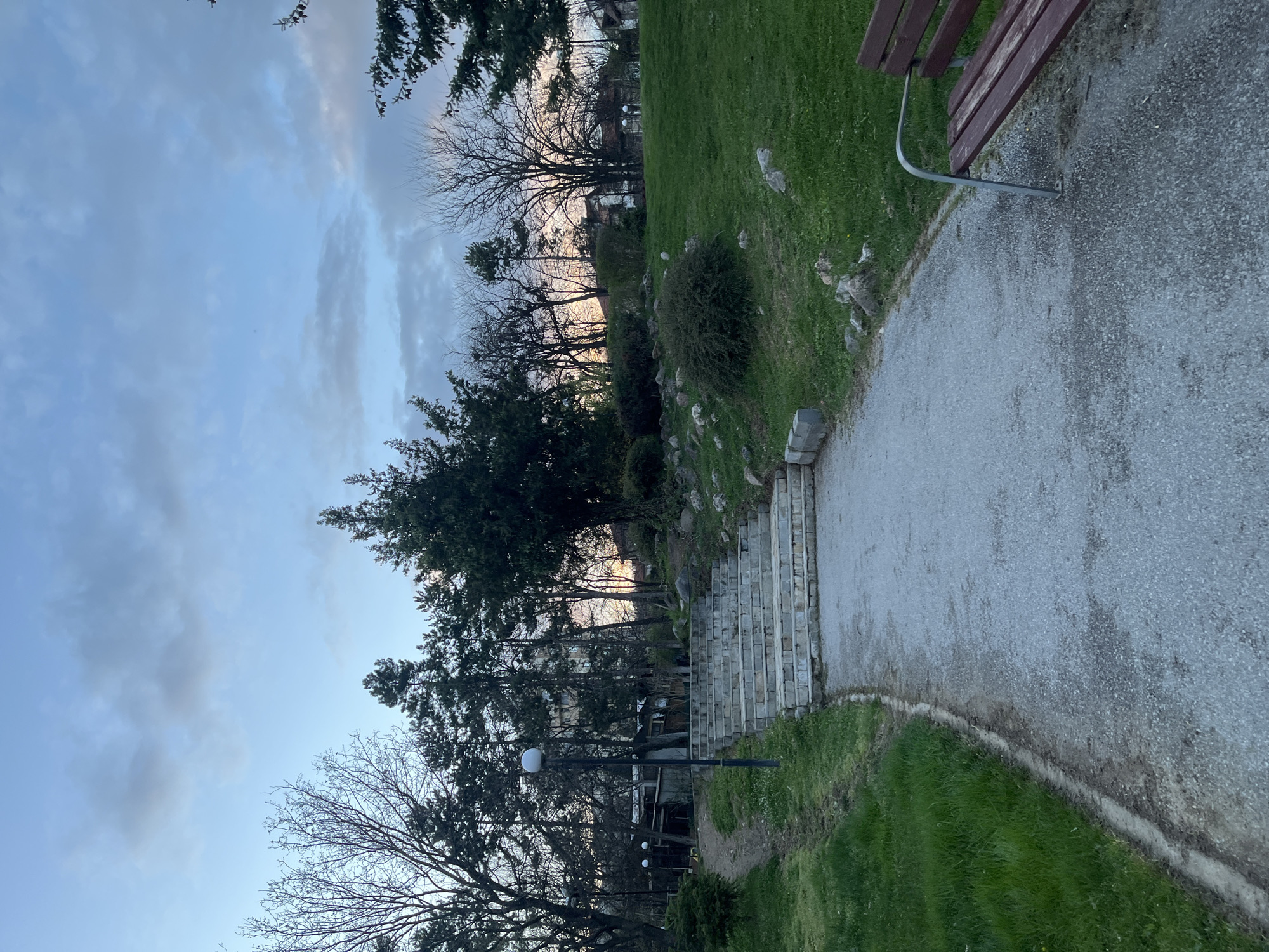 The image depicts a park setting. A gravel path runs along the right side of the frame, leading upwards. Adjacent to the path is a grassy area with trees and shrubs. A park bench is partially visible at the top right corner. Further up the path, stone steps can be seen leading to a higher elevation within the park. The sky is visible in the background, with a mix of blue and cloudy sections. The image is taken from a low angle. There are no people visible in the frame. The location is within the city of Goce Delcev, Bulgaria.