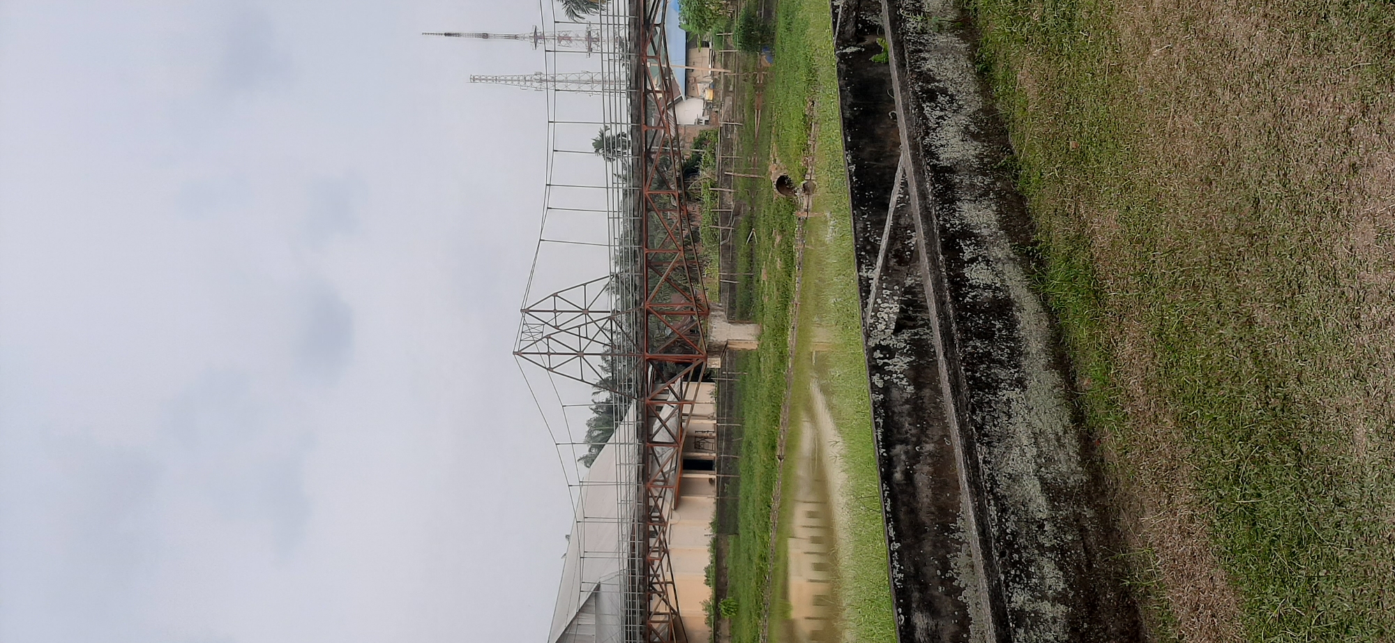 The image depicts an overcast day in Jambi, Indonesia, showcasing a partially constructed bridge spanning over a body of water. The bridge, made of rusty metal girders and cables, appears to be a suspension bridge with a central A-frame support structure. Adjacent to the bridge is a large, light-colored building with a metal roof, possibly a warehouse or industrial facility. The water in the foreground reflects the overcast sky and the building. On the bank of the water, a dark-colored bird or animal is visible standing near some grass. In the background, two tall radio or communication towers rise above the tree line. In the very foreground, there are concrete structures, possibly part of an unfinished drainage system or aquatic enclosure, with grass growing around them. The overall scene suggests an industrial or construction area near a waterway.