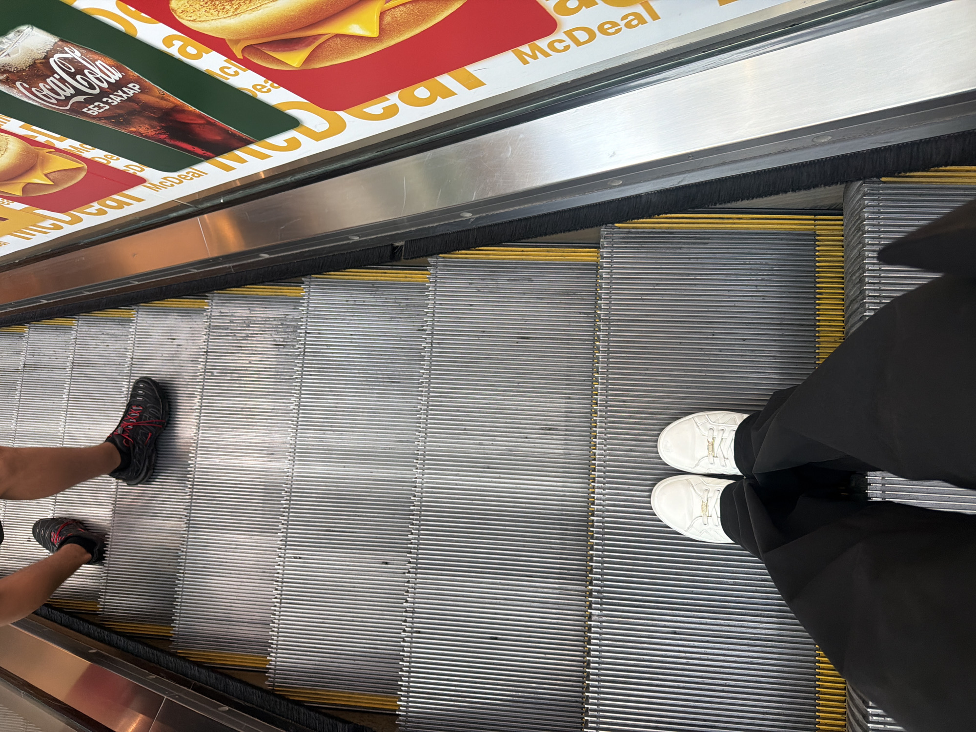 An overhead shot captures two people's legs and feet on an escalator. The escalator steps are metallic and grooved, with yellow safety lines. The person closer to the camera is wearing black pants and white sneakers. The person further up the escalator is wearing dark shorts and black athletic shoes with red laces. The escalator is bordered by a stainless steel railing. On the right side of the escalator, there are advertisements for McDonald's meals, featuring images of hamburgers and a Coca-Cola drink. The advertisements are predominantly red and yellow. The scene is indoors, likely in a shopping mall or similar public space.