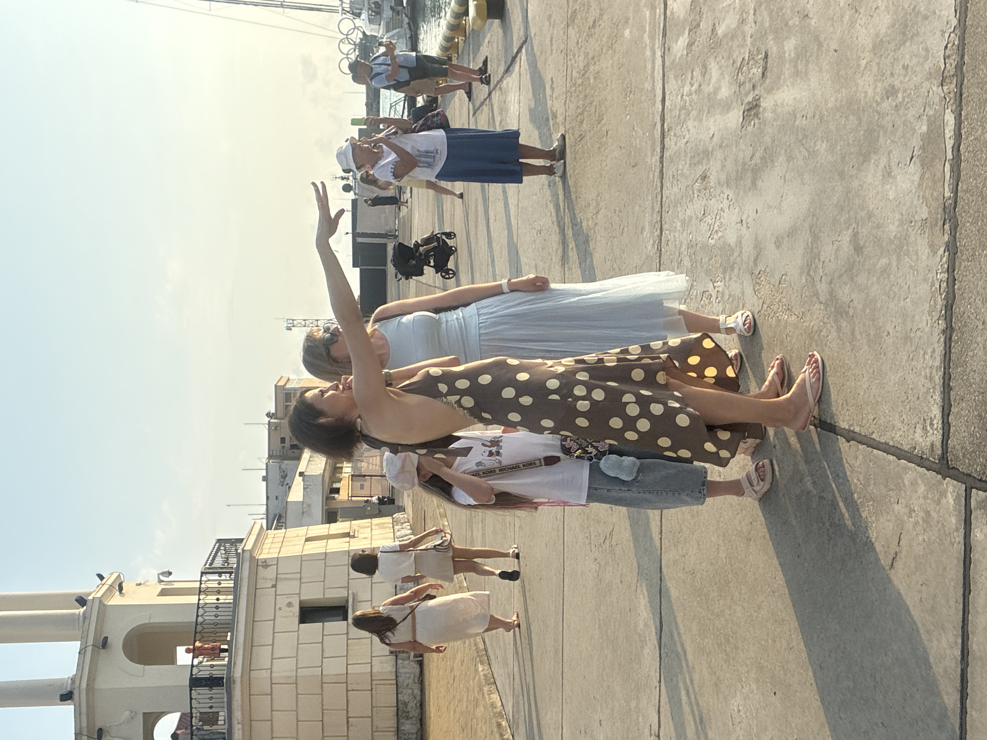 In Sochi, Russia, at the Sochi Sea Port, a group of people are gathered on a paved promenade under a clear sky with gentle sunlight. In the foreground, a woman in a polka-dotted dress is pointing towards the right, her arm extended, while a young girl in a white t-shirt and denim shorts stands beside her, looking in the same direction. To their left, two women walk away from the camera, one in a light-colored dress and the other in a light skirt and white top.
Further back, a woman in a light blue dress is looking towards the direction the first woman is pointing. To the right, a woman in a white hat and blue skirt is taking a photo with her phone, while a man in a blue t-shirt and shorts also holds a phone, as if capturing a moment. A stroller is visible behind the woman in the blue dress. In the background, a large sailing yacht with masts is docked in the harbor, and buildings of the port are visible. The overall scene suggests a relaxed atmosphere, with people enjoying the waterfront.