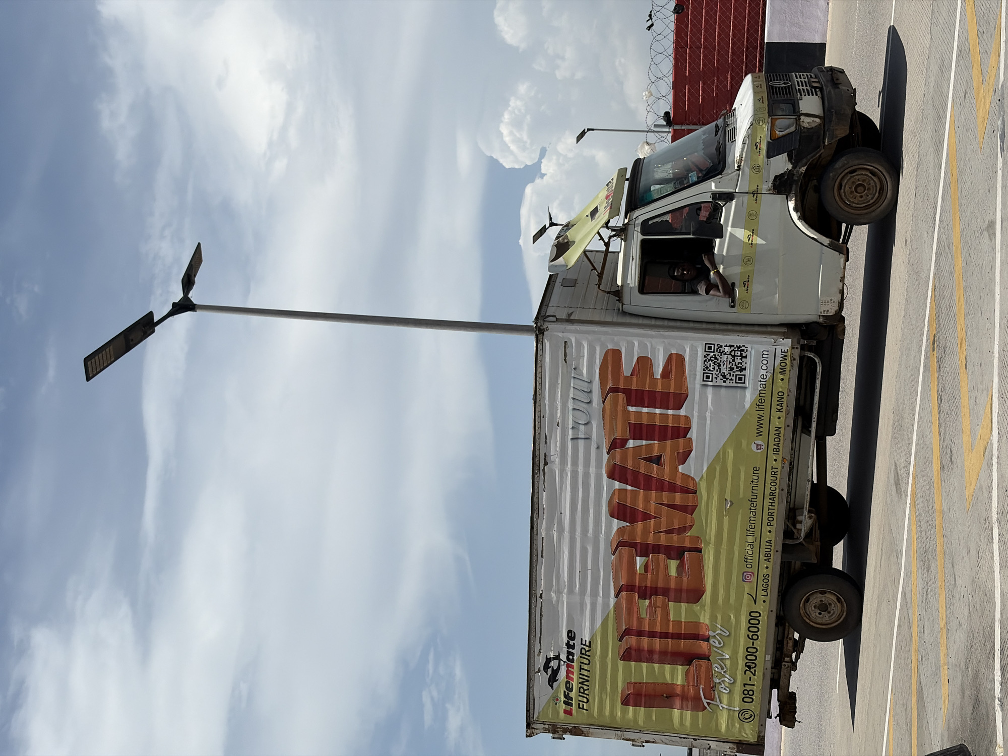 A commercial box truck with the brand "Lifemate FURNITURE" displayed on the side is parked on a light gray asphalt surface. Yellow lane lines are painted on the asphalt. The truck is oriented with its front facing the right side of the image and the rear facing the left side. A street light is positioned near the rear of the truck, extending into the cloudy sky. The background includes a fence topped with barbed wire, and a red wall behind it. There is a person in the truck. The image appears to be taken during daylight. The location is Igboefon, Nigeria.