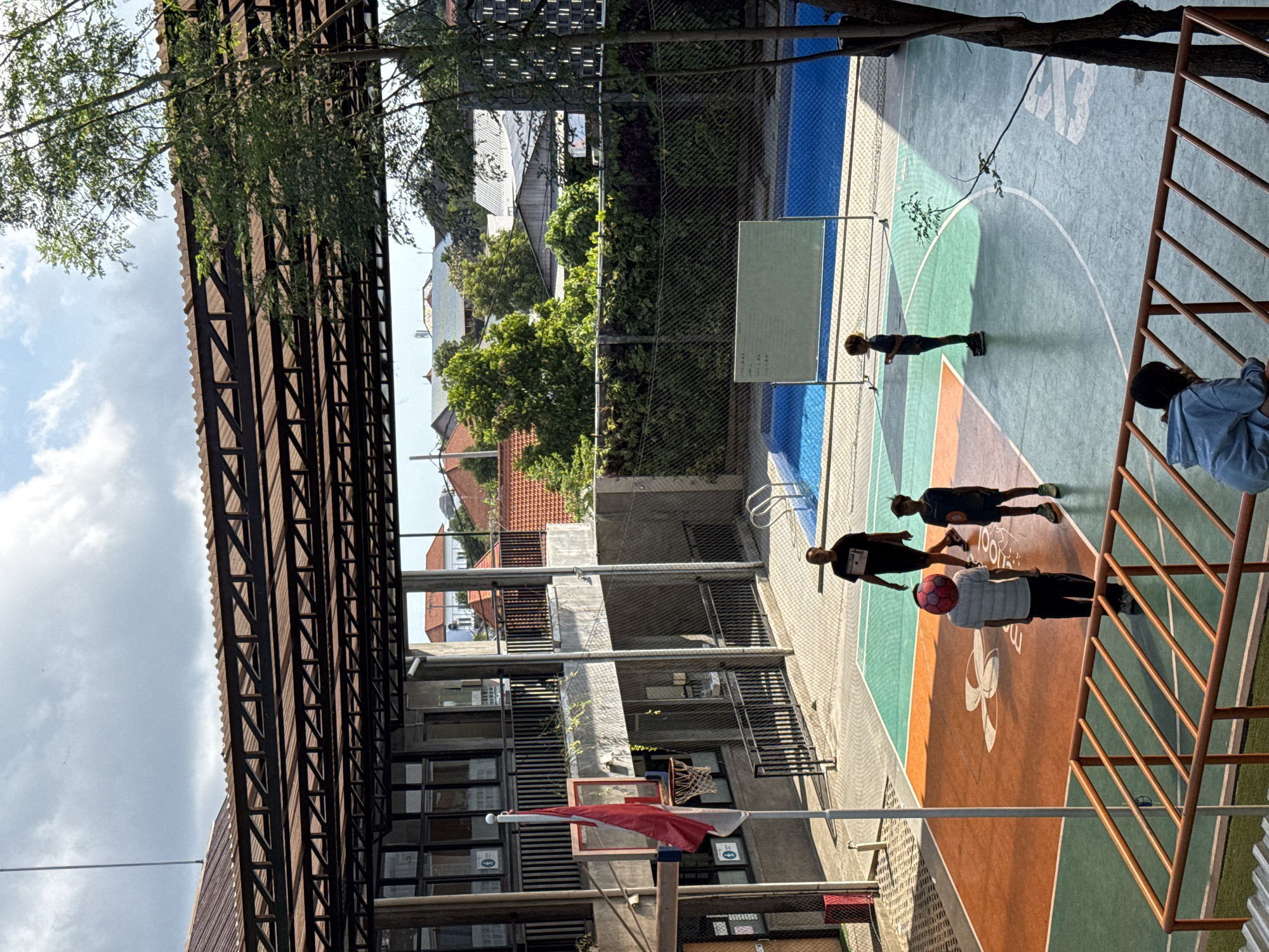 The image depicts an outdoor basketball court with a pool and a building in the background. The basketball court is multi-colored with orange, green, and light blue sections. There are four people visible on the court. One adult male is walking away from the camera, another adult male is standing near the basketball hoop with a red ball on his head, a young boy in shorts is standing and looking towards the basketball hoop, and another young boy is standing further away on the court. In the background, there is a swimming pool with blue water and a white whiteboard. The building in the background has multiple stories and balconies. The overall environment appears to be a school or a recreational facility located in North Kuta, Indonesia.