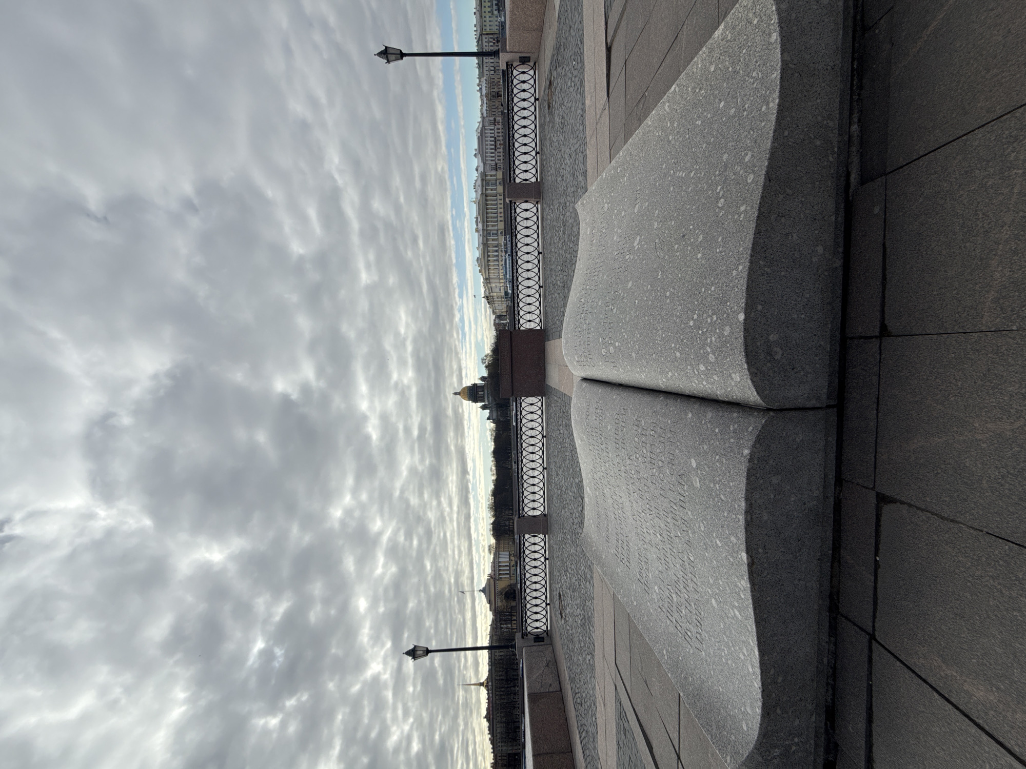 The image captures a scene along a granite embankment in Saint Petersburg, Russia. A large, light gray granite structure, possibly a bench or a part of the embankment itself, dominates the foreground. In the midground, a decorative metal fence runs parallel to the embankment, separating it from the water. Beyond the fence, the skyline of Saint Petersburg is visible, including recognizable landmarks such as the golden dome of Saint Isaac's Cathedral and the spire of the Peter and Paul Fortress. Street lamps are placed at intervals along the embankment. The sky is overcast with textured clouds.