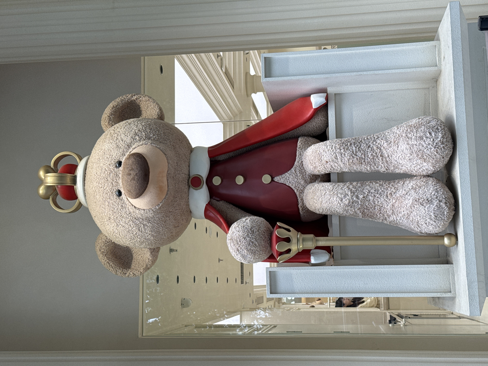 A sideways view of a large, fuzzy teddy bear dressed in a red royal coat with white trim and gold buttons, wearing a gold crown with a red gemstone, and holding a gold scepter with a decorative head. The bear is placed on a white, multi-tiered display stand within what appears to be a store window. The window reflection shows building elements and what could be people inside. The scene is in Jinjiang District, China.