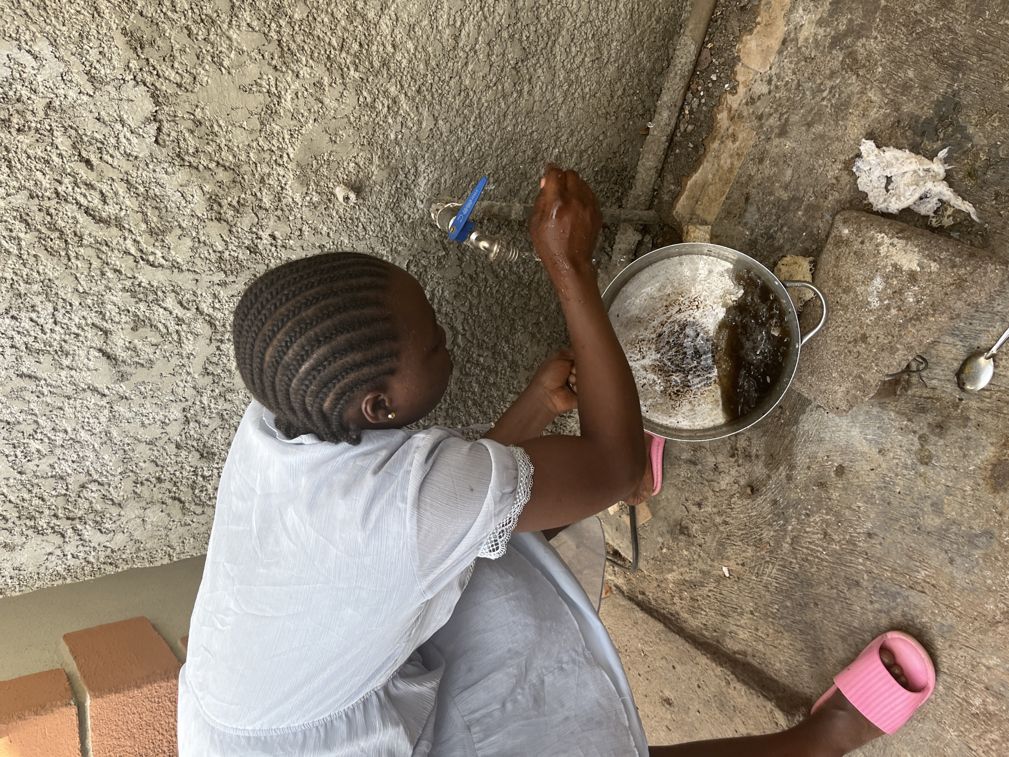 The image provides an overhead view of a person engaged in an outdoor or semi-outdoor activity involving water in Jos, Nigeria. The central subject, likely a woman or girl, is seen from behind, her back mostly to the camera. She has neatly braided dark hair and dark skin, and wears a light-colored, possibly white or light blue, short-sleeved dress with lace detailing on the sleeves. A small, subtle earring is visible on her left ear.
The person is operating a water faucet mounted on a rough, textured concrete wall. Her hands appear wet, indicating she is actively using the water. The faucet features a blue handle with "RBM ITALY" visible on it. Below the faucet, a metal pot or basin, appearing to be made of aluminum, rests on the ground. It contains dark, murky water, with some residue visible along its inner surface.
The ground is a mix of concrete and possibly packed earth, uneven and showing signs of wear and dampness, especially around the water source. To the right of the pot, a silver spoon lies on the ground, near some crumpled white material, possibly waste. In the lower-left corner, parts of reddish-brown blocks, resembling bricks, are visible. The scene suggests a daily chore involving water collection or washing. The lighting indicates it is daytime, likely under an overcast sky or in a shaded area, providing soft illumination. The overall setting is functional and utilitarian.