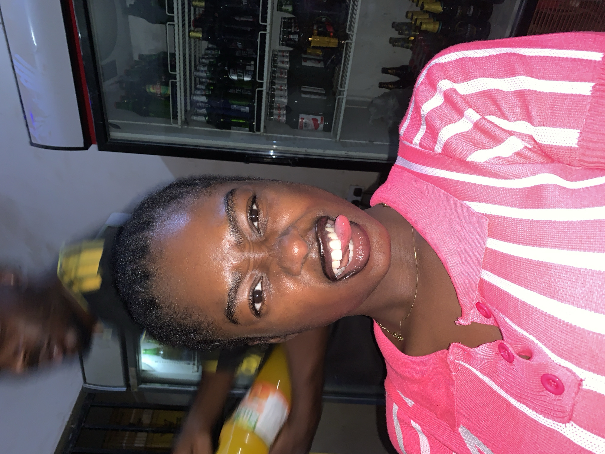 A woman with dark skin and short hair is sticking her tongue out at the camera. She is wearing a pink and white striped shirt and a gold necklace. She is standing in front of a refrigerator filled with bottles and cans. A man with dark skin is to her left holding a yellow bottle. The location is likely New Stadium, Nigeria.