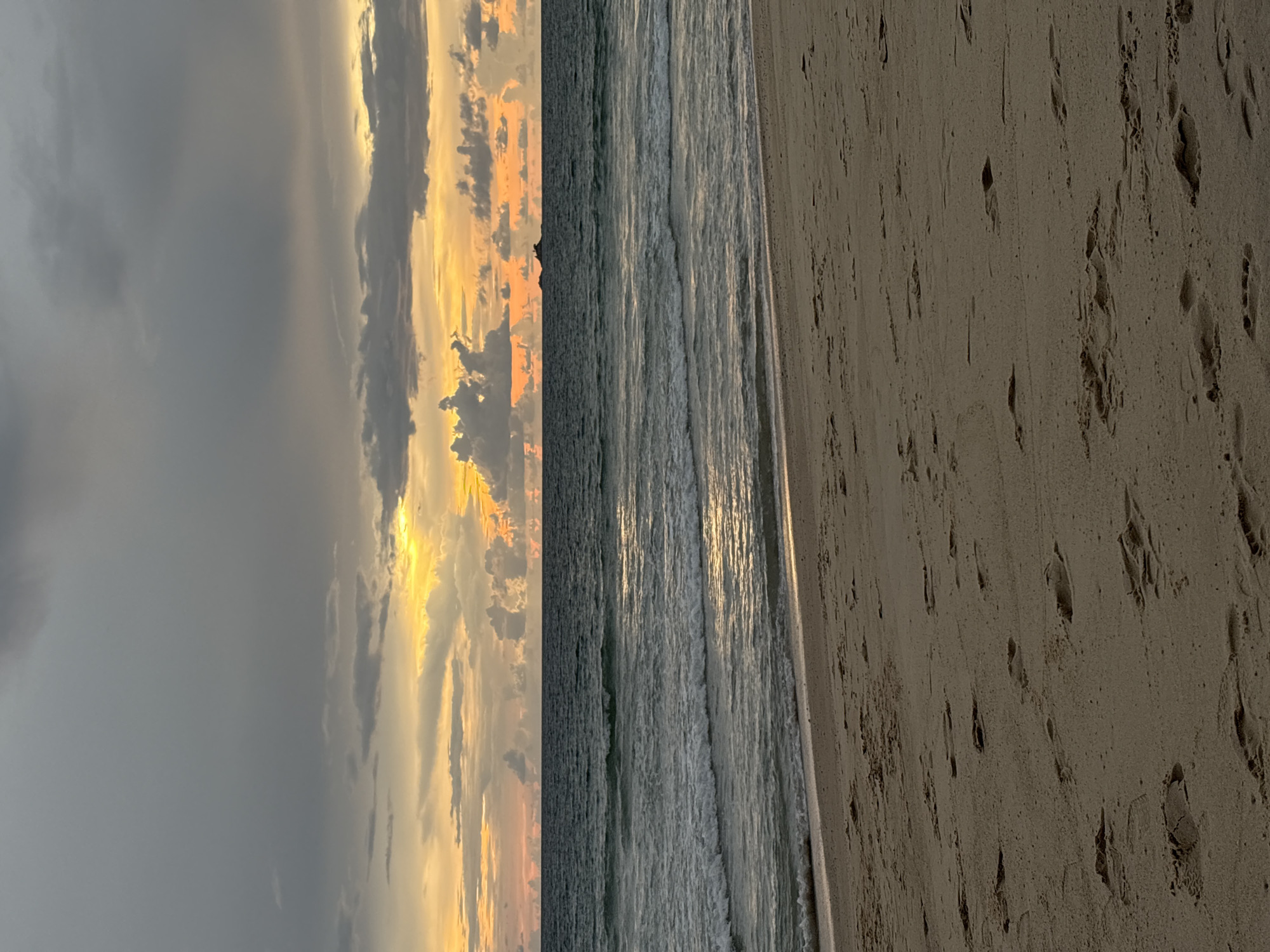 This is a photograph of a beach scene in Choeng Thale, Thailand, likely taken during sunset or sunrise. The image is oriented vertically, with the sky on the left, the ocean in the middle, and the sandy beach on the right.
The sky is filled with dramatic clouds. The left side of the sky is dominated by dark, heavy gray clouds, suggesting an approaching storm or the aftermath of one. However, towards the right, the clouds are illuminated by the sun, creating a vibrant display of orange, yellow, and pink hues. The sunlight breaks through the clouds in streaks, casting a warm glow.
The ocean is a deep, textured gray with visible waves rolling towards the shore. The water reflects the colors of the sky, creating shimmering lines of light on the surface. The waves appear to be moderately sized, with white foam cresting.
The foreground is a sandy beach, with the sand appearing light brown and textured. Numerous footprints are visible in the sand, suggesting recent human activity, though no people are directly depicted in the image. The footprints are distinct and varied in size and depth.
The time of day is clearly indicated by the dramatic lighting in the sky, characteristic of either dawn or dusk. The weather appears to be a mix of overcast and bright conditions, with the sun breaking through. The location cues are minimal, with the prompt providing the specific location. The overall mood of the image is serene and evocative, blending the raw power of nature with the subtle traces of human presence. No visible text is present in the image.
