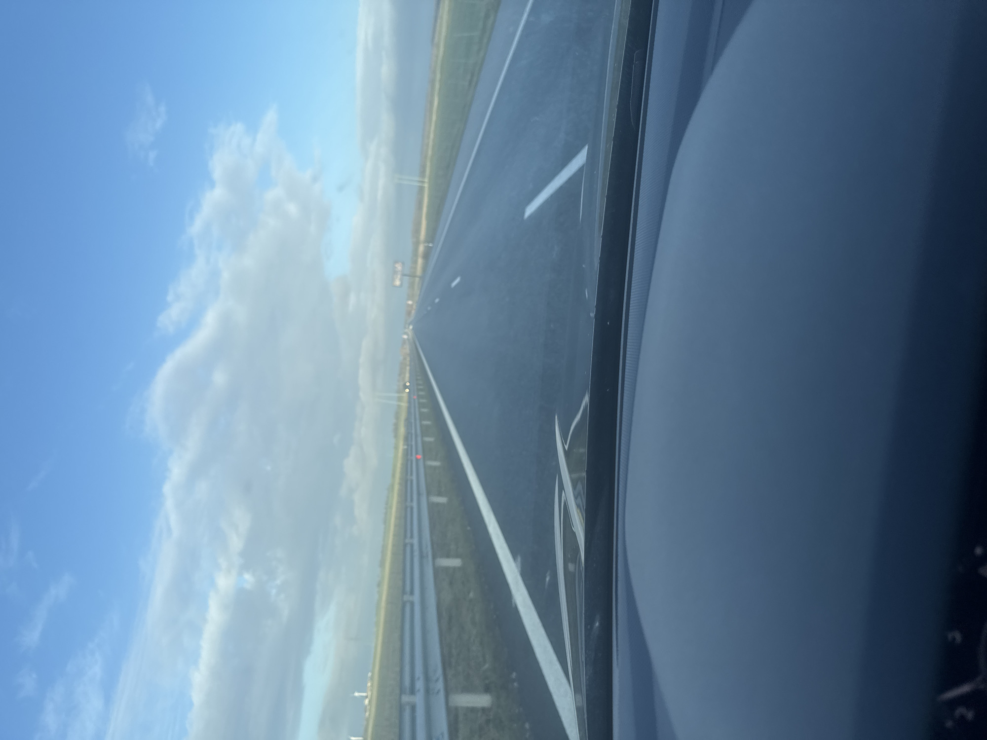 The image depicts a forward-facing perspective from the interior of a vehicle on a multi-lane highway in the city of Палаузово, Bulgaria. The dark dashboard of the vehicle is visible across the bottom of the frame. The asphalt roadway extends straight into the distance, featuring a solid white line marking the left shoulder and a dashed white line indicating a lane division. To the left of the highway, a segmented silver metal guardrail runs parallel to the road, beyond which is a grassy embankment. In the far distance on the left horizon, several white wind turbines are discernible. The right side of the highway is bordered by a flat, grassy area. The sky above is partially clear blue with scattered white and grey clouds. Ambient daylight conditions prevail. No individuals or active interactions are visible.