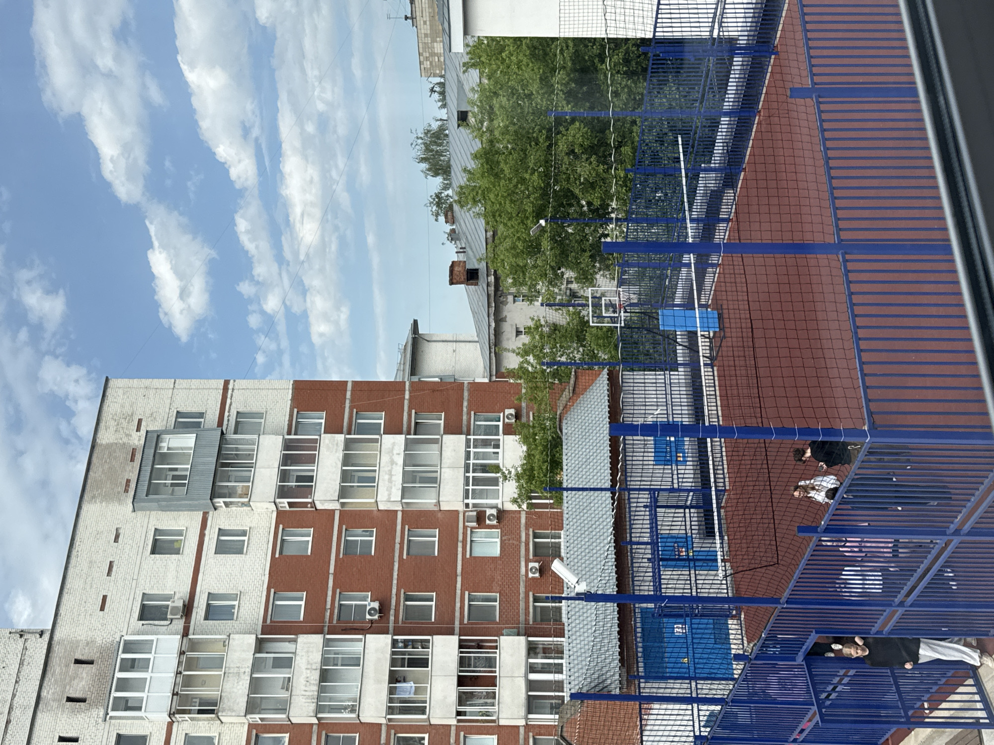 The image shows a view from an elevated position, likely a balcony or window, overlooking a courtyard or playground in Perm, Russia. A tall, multi-story building, likely a residential apartment block, is visible on the left, with a mix of white and red brick facade and numerous windows. Air conditioning units are mounted on the building's exterior.
In the foreground, there is a blue metal fence or structure of some sort, possibly part of the playground or sports area. The ground within the enclosure appears to be a red or brown material. Several people are within the enclosed space, seemingly interacting with each other.
In the middle ground, a basketball hoop is visible, partially obscured by the fence. Above the building, the sky is bright blue with scattered white clouds. Trees are visible in the background, adding to the urban landscape.