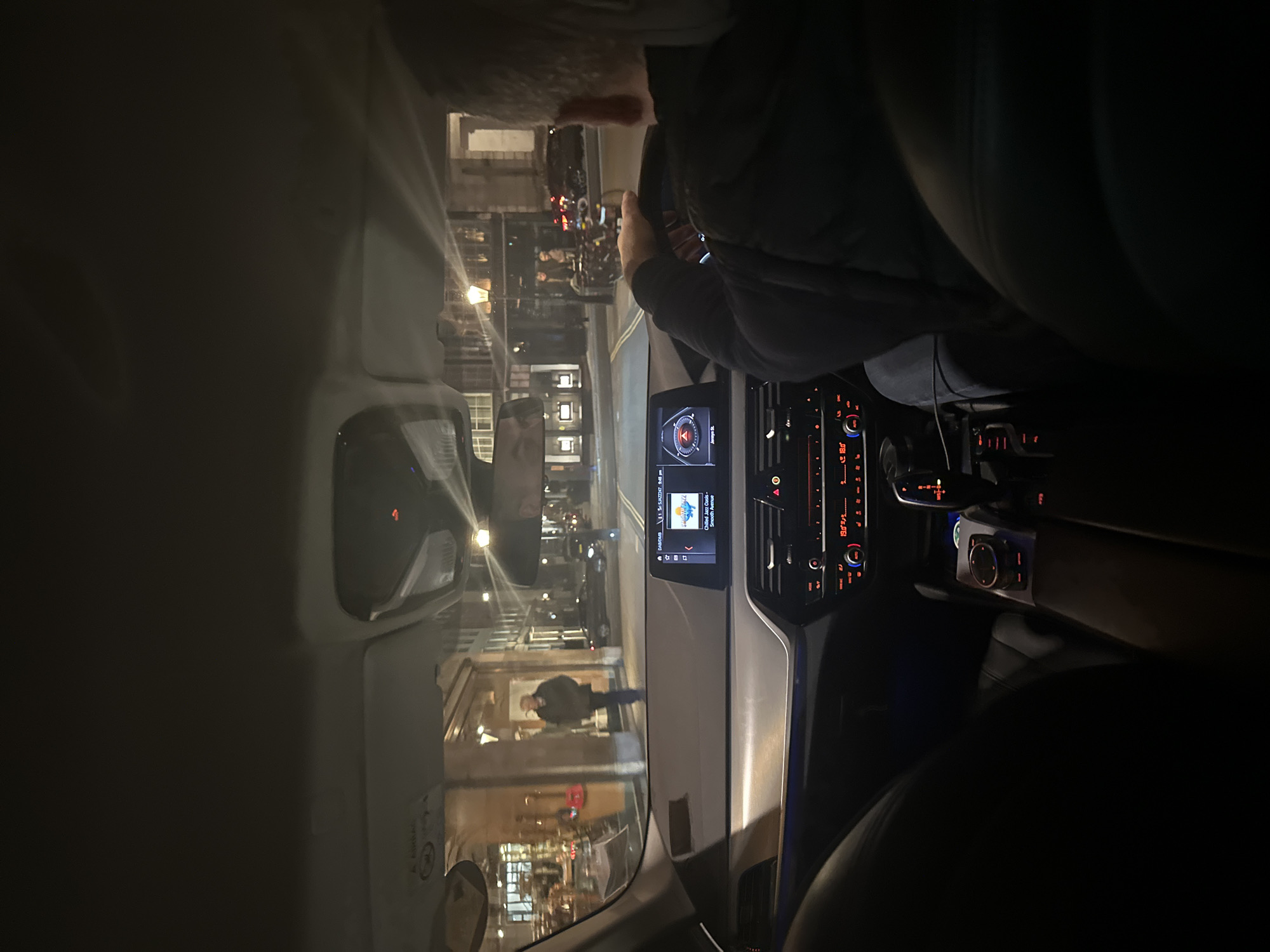 The scene is captured from inside a right-hand drive vehicle, likely a modern BMW, at night. The location is Jermyn Street, St James's, London, United Kingdom, identified by street signage ("Jermyn St.") on the vehicle's infotainment system.
A male driver, with grey hair, is visible from behind in the foreground right, gripping the steering wheel. He wears a dark, padded jacket. The vehicle's dashboard features a central infotainment display showing navigation information and entertainment details ("Chilled Jazz Groovs - Smooth Avenue"). Climate controls below indicate a temperature setting of 19.0°C (or 18.0°C). An electronic gear selector and iDrive controller are present on the center console.
Outside, a paved street extends forward, lined by multi-story commercial buildings with illuminated shopfronts. A single male pedestrian, wearing a dark jacket and trousers, walks on the left-side pavement, moving away from the vehicle. Traditional-style streetlights illuminate the scene. In the mid-ground, other parked vehicles and a bicycle rack are visible on the right side of the street. The environment depicts an urban commercial street at night.