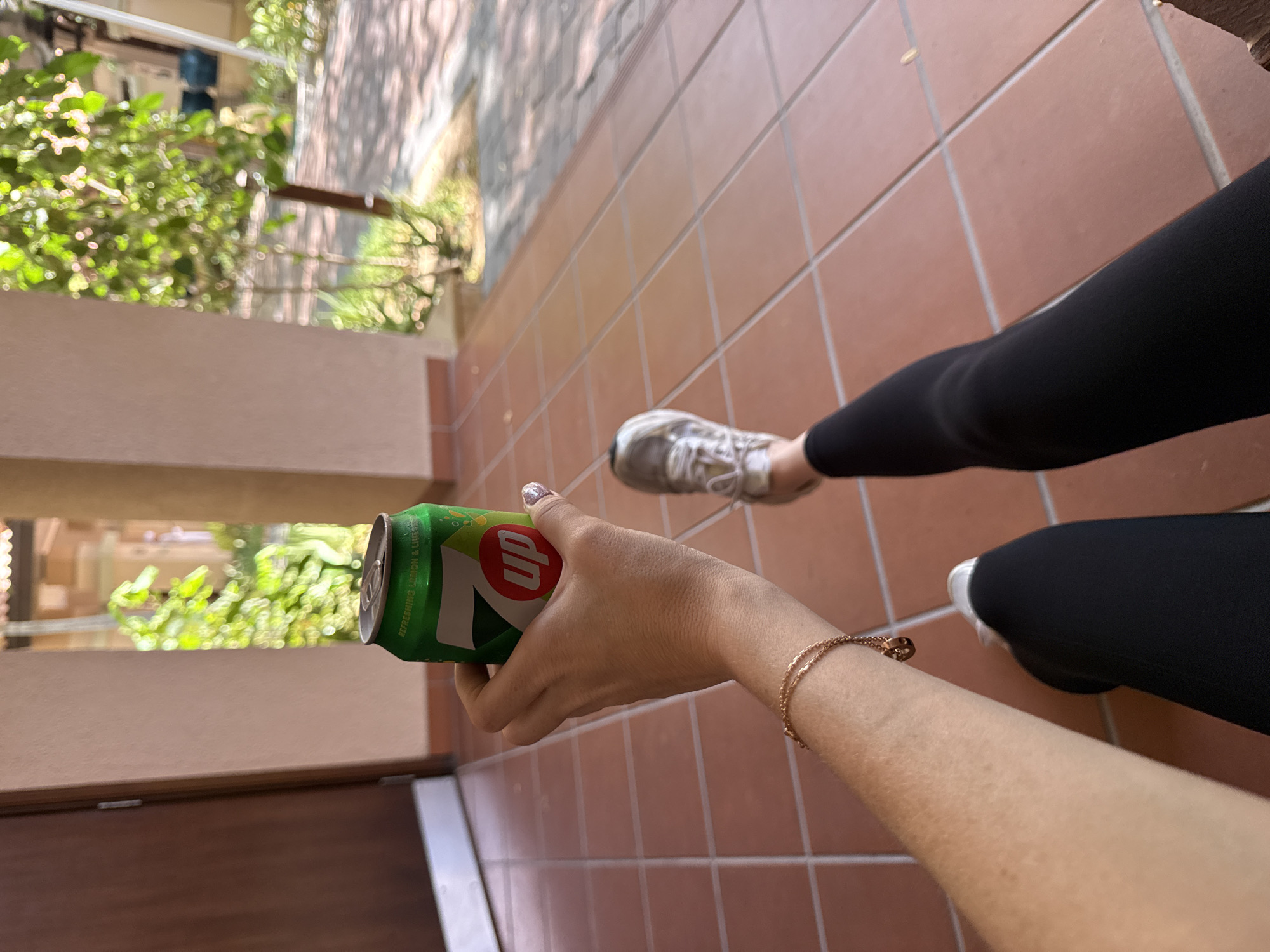 A first-person perspective photograph shows the lower body and right arm of an individual. The individual's right hand holds a green 7 Up soda can, displaying the "7up" logo and text "REFRESHING LEMON & LIME." A delicate, rose gold-colored double-chain bracelet with a small circular charm is visible on the right wrist. The fingernails of the right hand appear to have light-colored, possibly glittery, polish. The individual's legs are extended, clad in plain black leggings. Light-colored (likely white or very light grey) athletic sneakers with white laces are worn on the feet. The individual is situated on a paved surface composed of square, terracotta-colored tiles with visible grout lines. To the left, a brown wooden door frame and a light-colored wall are visible. To the right and in the background, lush green foliage, an outdoor paved area, and shadows indicate bright daylight conditions. Further in the background, an out-of-focus structure with a dark blue decorative pot is faintly discernible. There are no definitive visual markers within the image to confirm the specific location as Dubai, United Arab Emirates.