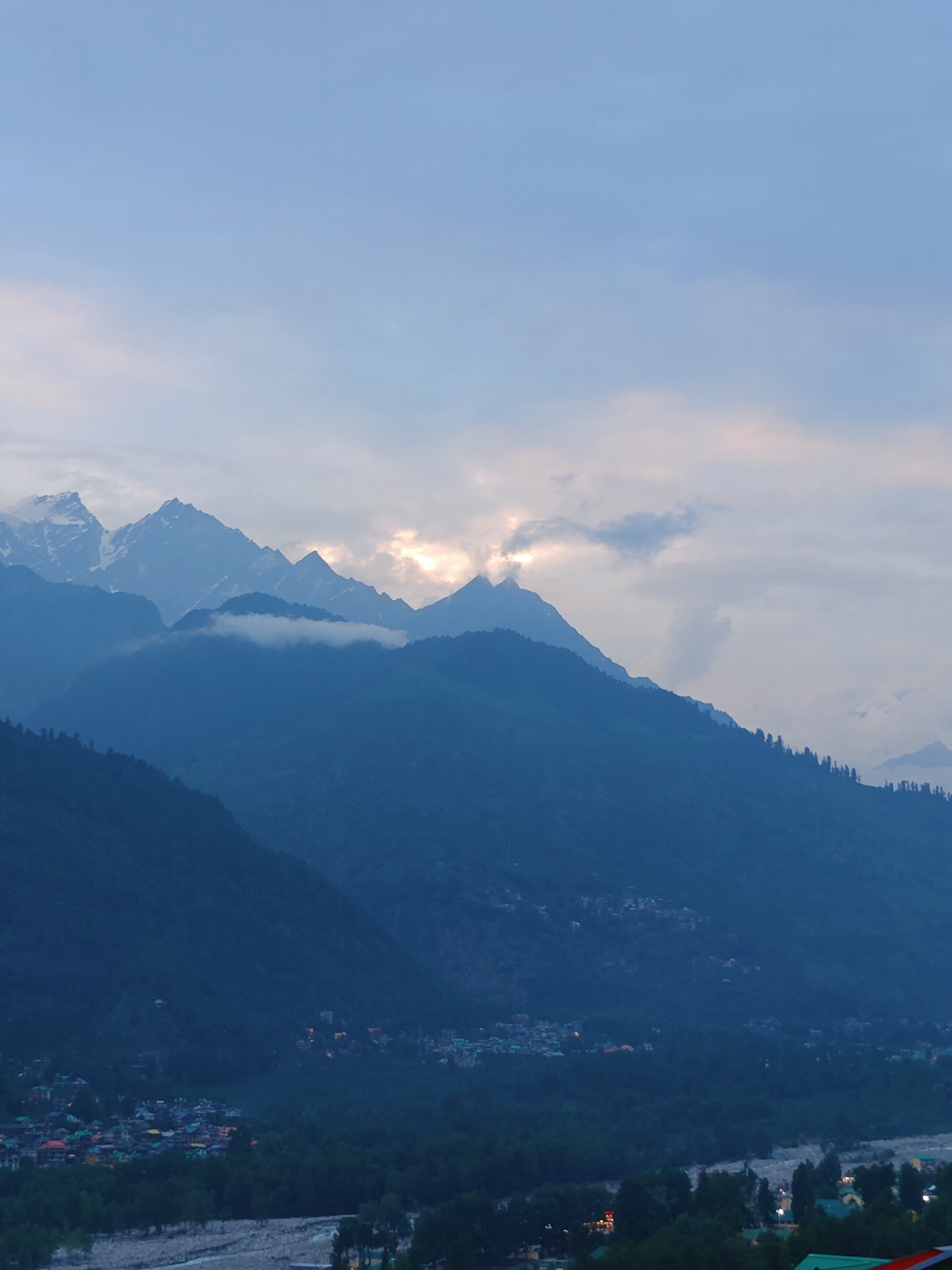 The image depicts a panoramic view of the mountainous landscape surrounding Vashisht, India, likely taken during dawn or dusk given the soft lighting and muted colors. Majestic, layered mountain ranges dominate the scene, with snow-capped peaks visible in the distance, suggesting a high-altitude location. Wisps of clouds are draped across the mountains, adding depth and texture. In the foreground, a valley floor is visible, dotted with a settlement. The settlement appears to be a small town or village, with numerous buildings exhibiting colorful roofs, indicative of residential areas. A riverbed, likely a shallow stream or river, is present in the lower portion of the valley, its presence suggested by a rocky, uneven terrain. Dense green foliage, characteristic of a forested area, covers the slopes of the mountains and the valley floor, interspersed with the built environment. The overall atmosphere is serene and tranquil, highlighting the natural beauty of the Himalayan region.