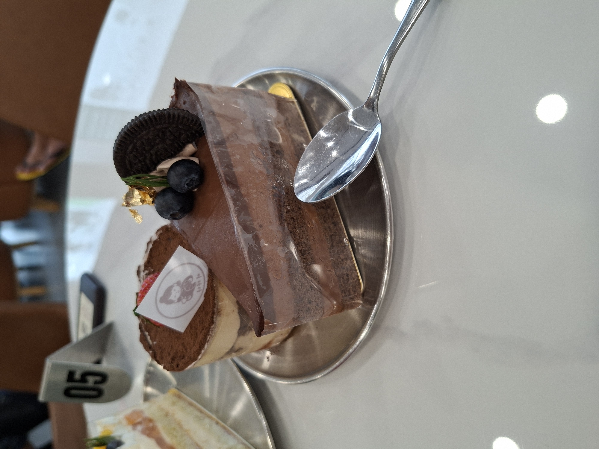 A high-angle shot shows a plated dessert consisting of a chocolate cake slice wrapped in clear plastic and a cylindrical cake roll topped with a logo-bearing sticker. Garnishments include blueberries, a halved Oreo cookie, and gold leaf flakes. A silver spoon rests on the plate next to the slice. A table number '05' is visible in the lower left. A second plate with a partially visible sandwich is present. The scene is set in Nha Trang Ward, Vietnam.