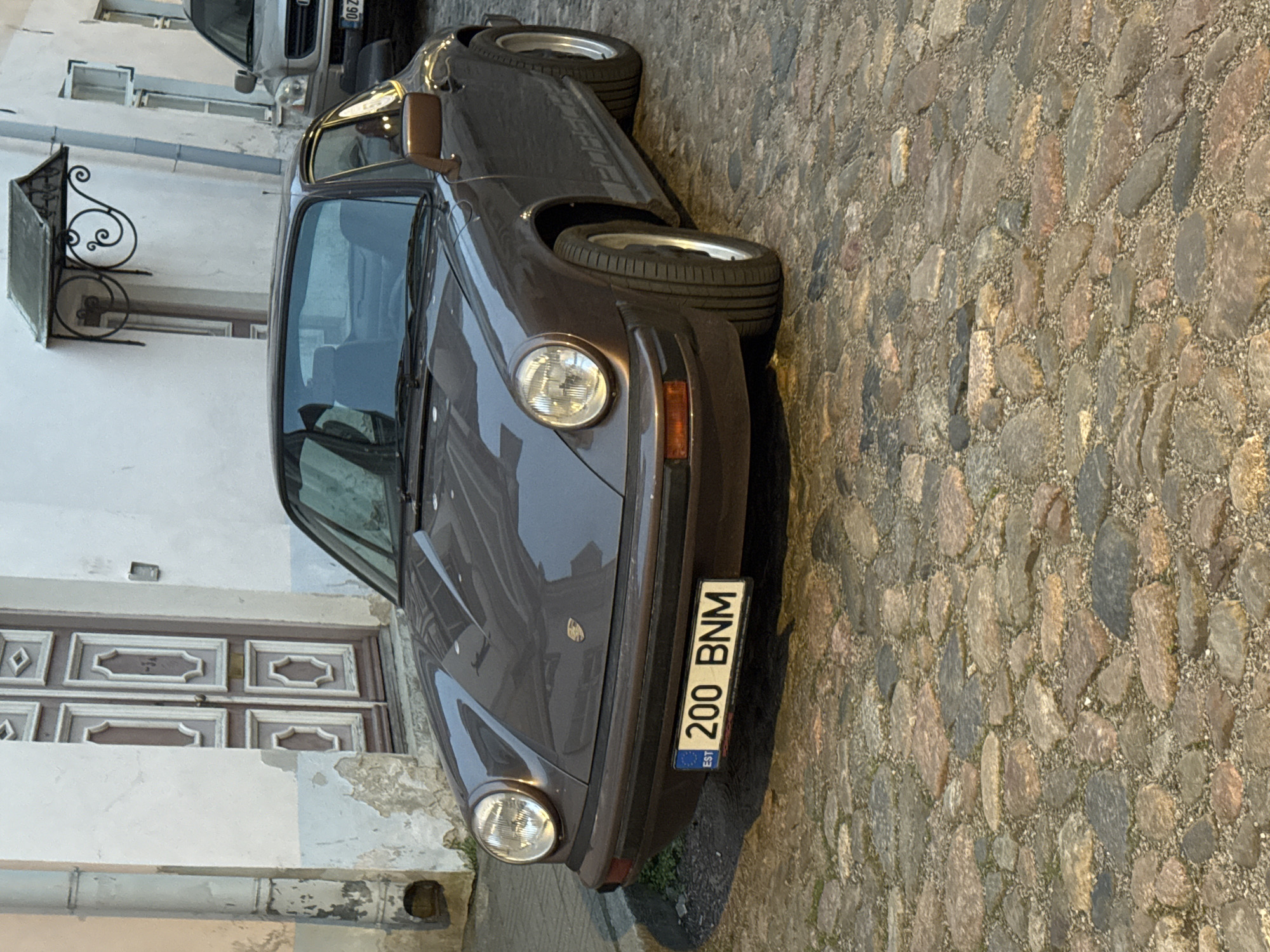 A brown Porsche 911 is parked on a cobblestone street in Tallinn, Estonia. The car has a license plate that reads "200 BNM" and features the EST symbol. The background shows a white building with decorative doors and a street lamp. Another vehicle is visible parked in the background. The scene appears to be an exterior shot during daylight.