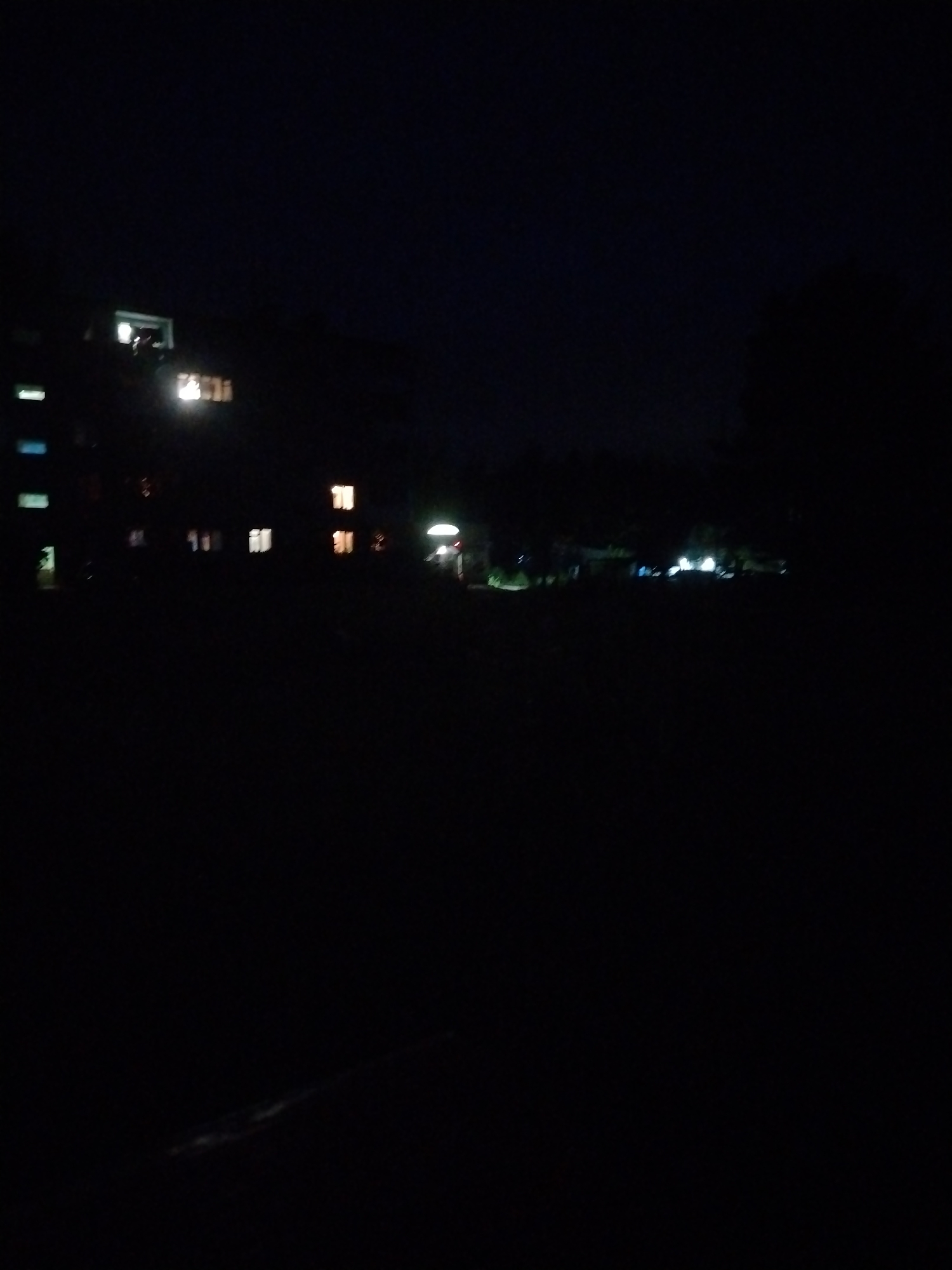 This is a nighttime photograph taken in the city of Stroitel', Russia. The scene depicts an apartment building with several illuminated windows, suggesting occupancy. The building appears to be a multi-story structure, common in urban environments. In the distance, there are other faint lights, possibly from other buildings or streetlights, indicating the presence of a developed area. The foreground is largely obscured by darkness, making it difficult to discern specific details of the immediate surroundings. The overall impression is a quiet, nocturnal urban landscape.
