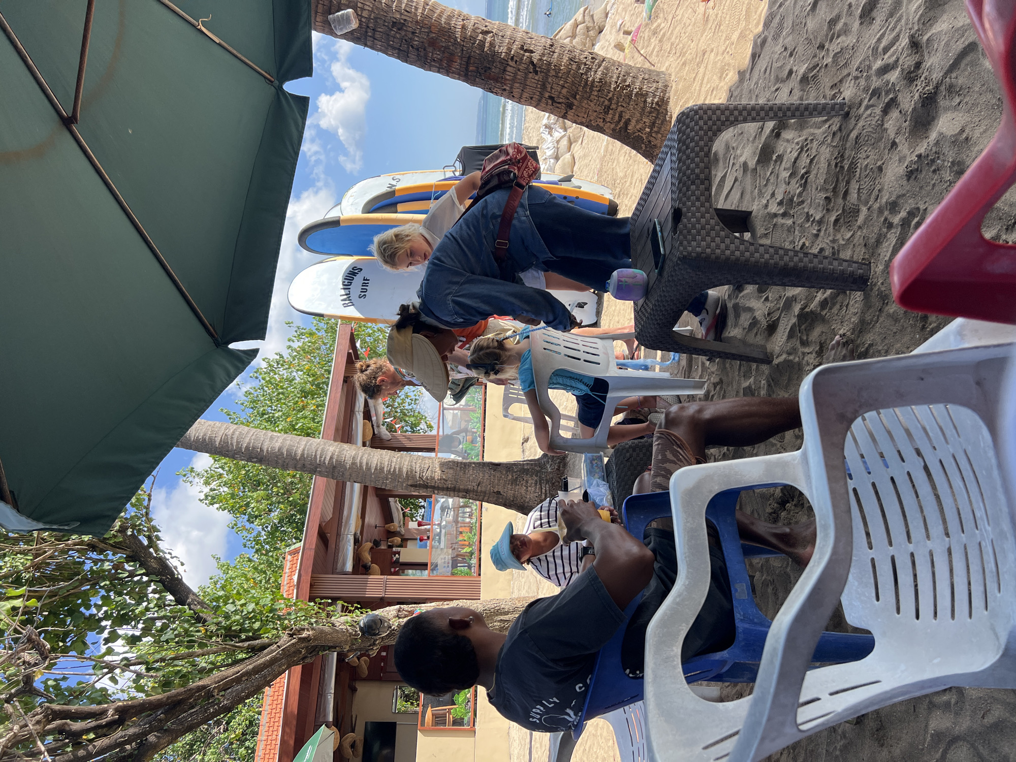 This image captures a group of people relaxing on a sandy beach, likely in Kuta, Indonesia, given the presence of surfboards. In the foreground, two individuals are seated in plastic chairs, one in a blue chair and another in a white one. The person in the blue chair, a dark-skinned male wearing a dark t-shirt, holds a drink. The person in the white chair is mostly obscured. Further back, a woman in a denim jacket and a straw hat is interacting with another person, possibly a surfer. Behind them, several surfboards stand upright. A large green umbrella provides shade. The background shows palm trees, a building with a reddish roof, and the ocean with gentle waves. The overall scene depicts a casual beachside atmosphere, possibly a surf school or rental spot.