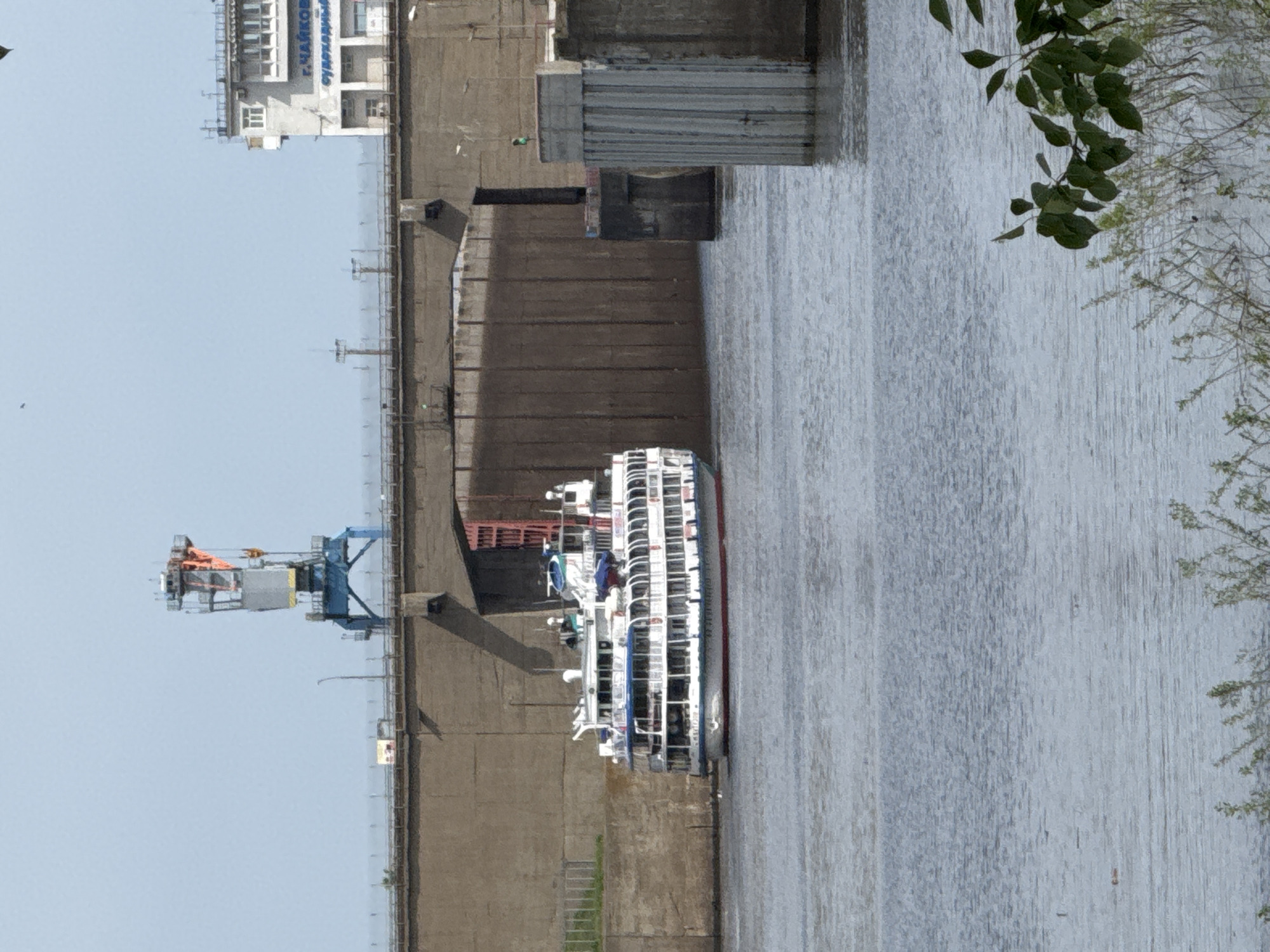 The image depicts a river scene in Chaikovsky, Russia, featuring a passenger ship passing through a lock. A large concrete structure forms the lock walls. On the left side, a crane is visible. A building with signage in Russian is situated further up. The river fills the right side of the frame, with foliage along the edge.