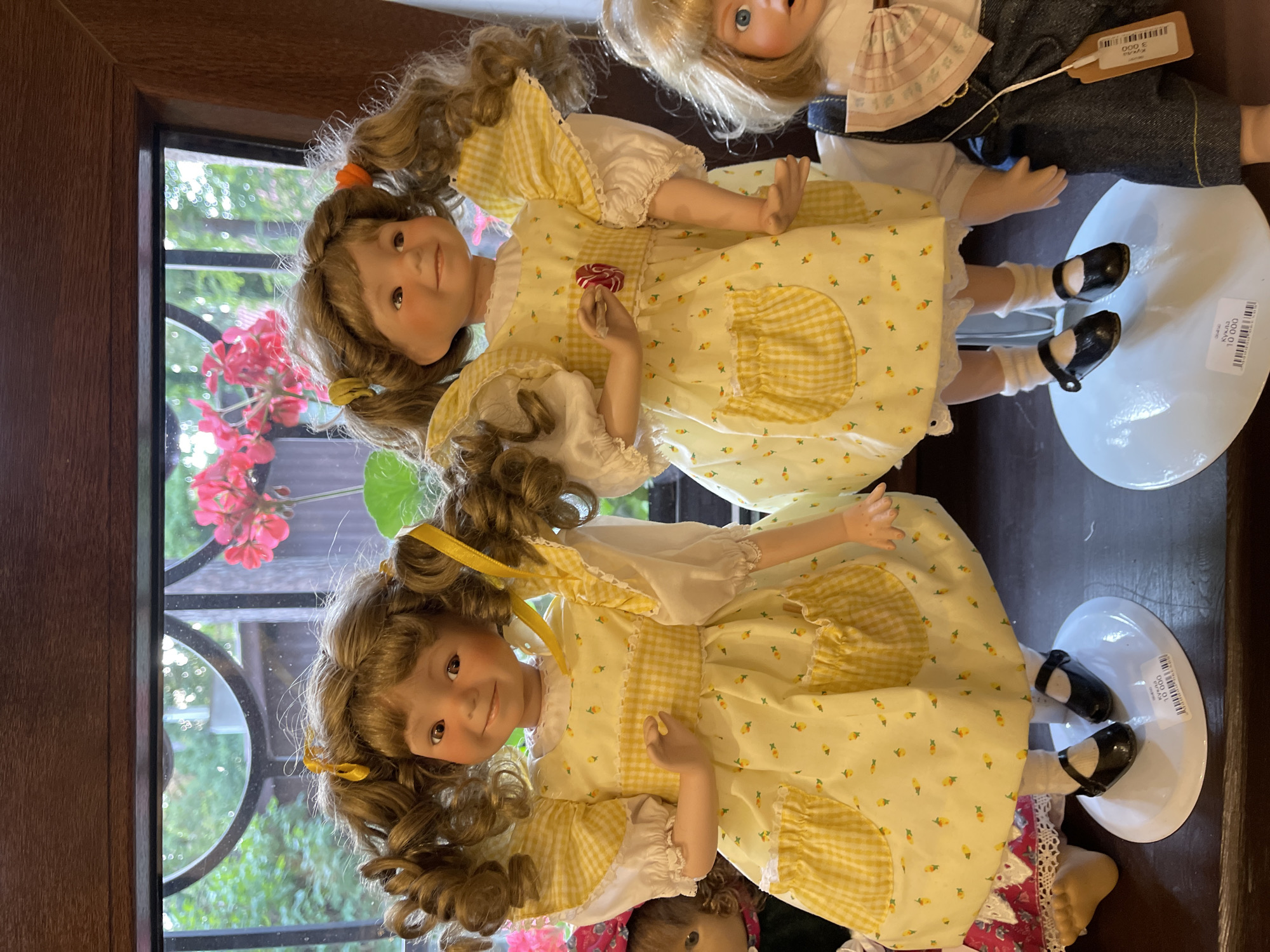 This is a close-up, eye-level photograph of two porcelain dolls, likely collectibles, displayed on white oval stands against a dark wooden surface. The dolls are positioned in the foreground, with a blurred background suggesting a window with greenery and pink flowers, hinting at an outdoor setting visible through glass.
The doll on the left is the primary subject, facing slightly to the right. She has curly, light brown hair pulled into two pigtails with yellow ribbons. Her face is round with rosy cheeks, brown eyes, and a smiling expression. She wears a yellow dress with a white collar and puffed sleeves. The dress features a white background with a pattern of small yellow fruits, a yellow and white gingham trim at the waist, and two front pockets also in gingham. A red and white swirl lollipop is affixed to the center of her chest. White socks and black mary jane shoes complete her attire. A white tag with black text is attached to the base of her stand.
To the right of the main doll, another doll is partially visible, standing on a similar white stand. This doll has blonde hair and blue eyes. She is wearing a white shirt and dark denim overalls with yellow stitching. A light pink bow with a decorative pattern is tied around her neck. An orange tag with black text is attached to the overall strap.
The overall scene suggests a display of collectible dolls in a shop or private collection, possibly in Svetlogorsk, Russia, given the inclusion of Russian text on one of the tags. The lighting is even and diffused, highlighting the details of the dolls and their clothing.
