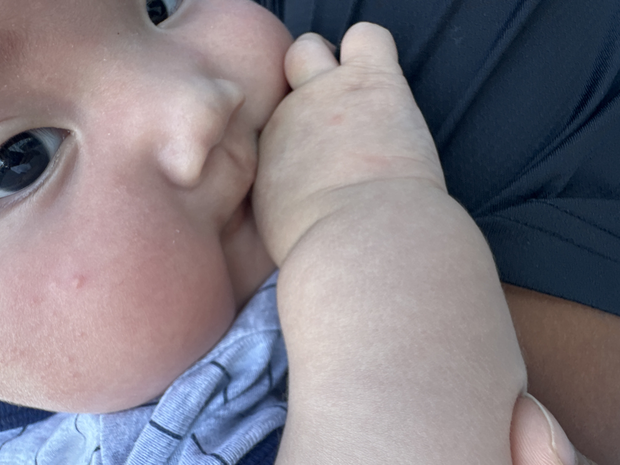The image is a close-up shot of a baby's face and arm. The baby has fair skin and dark eyes. The baby's left arm is bent and its hand is near its mouth, with fingers partially in its mouth. The baby is wearing a light blue striped shirt. In the background, a dark blue fabric with a textured pattern is visible, possibly clothing. The lighting suggests it could be daytime, but there are no specific cues about the time of day or weather. The setting appears to be indoors or in a shaded area. The overall mood is intimate and focuses on the baby's features and actions. No text is visible in the image.