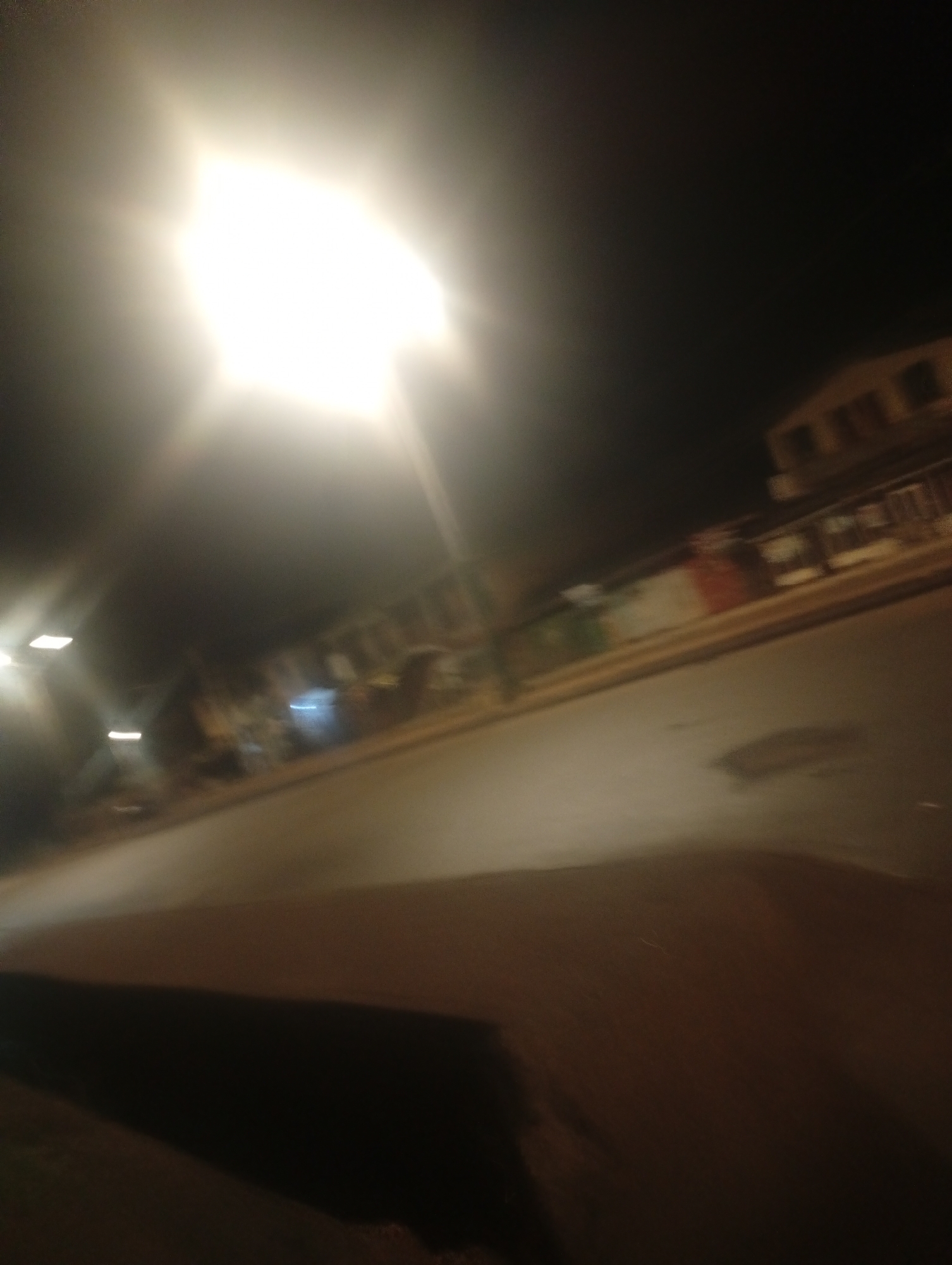 The image depicts a street scene at night in Bukuru, Nigeria. A bright, possibly street lamp, dominates the upper left of the frame, creating a lens flare and casting a strong glow that washes out details. In the background, blurred structures of buildings are visible along the street. One building on the right appears to be a two-story structure with visible windows. The street itself is dark asphalt, with a slight sheen suggesting it might be wet, or simply reflecting the ambient light.
There are no clearly identifiable people or specific objects in the foreground or mid-ground due to the motion blur and low light conditions. The overall impression is that of a quiet street at night, possibly illuminated by artificial lighting. The dark sky and the bright light source suggest it is nighttime. There is no visible text in the image. The blurry nature of the image suggests it was taken with a moving camera, possibly from a vehicle.