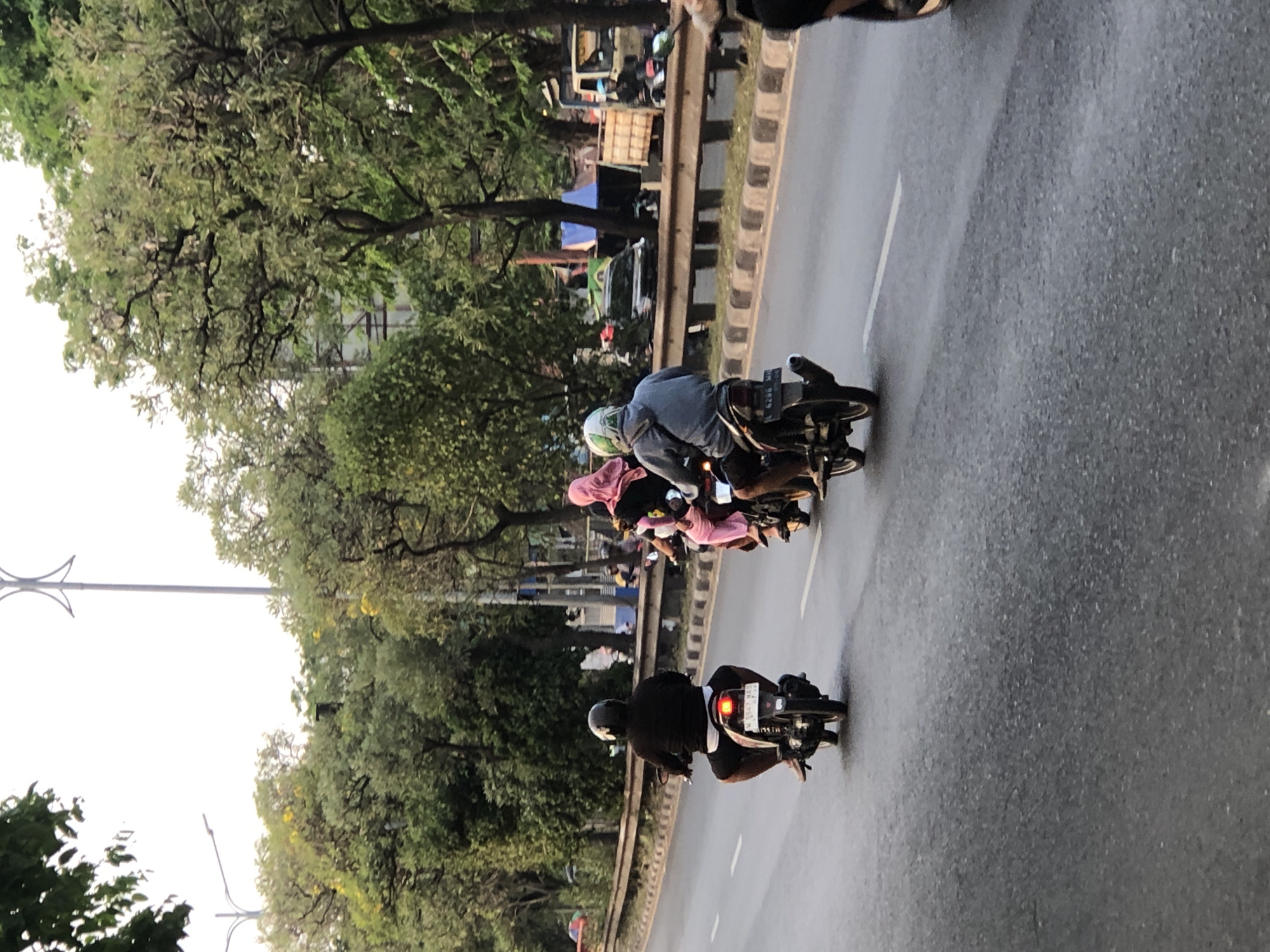 The image shows a street scene in Kludan, Indonesia, with three motorcycles riding on a paved road. The foremost motorcycle has a rider wearing a black long-sleeved shirt and white shorts, with a black helmet. Behind them is another motorcycle with a rider wearing a pink hijab and black pants, followed by a third motorcycle with a rider wearing a grey hooded jacket and shorts, and a green and white helmet. The road is bordered by a metal guardrail and a sidewalk with trees and vegetation behind it. In the background, there are more trees, some buildings, and a large truck. The sky is overcast, suggesting it might be late afternoon or early morning. The overall scene depicts everyday traffic and urban life in Kludan.