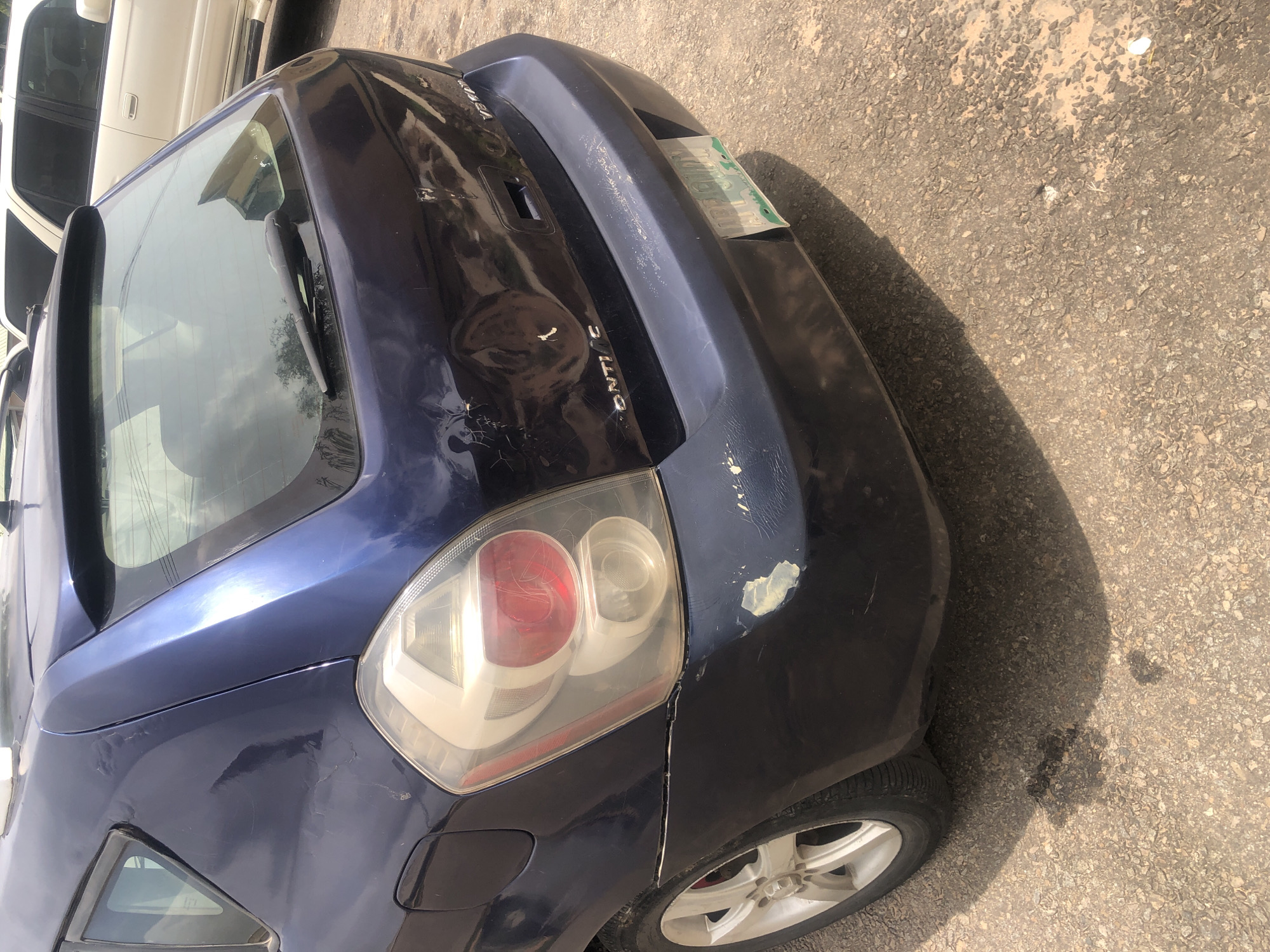 The image shows the rear left side of a dark blue hatchback car, likely a Pontiac Vibe, parked on a paved road. The car has visible damage, including dents and paint chips on the rear bumper and quarter panel. The taillight assembly on the left side is intact but appears scuffed. A license plate is partially visible on the rear bumper. The car is parked outdoors on a sunny day, as indicated by the shadows. The surrounding environment appears to be a street or parking area in Jalingo, Nigeria, with cracked asphalt and some debris on the ground. In the background, a light-colored SUV is partially visible. No people are present in the image.