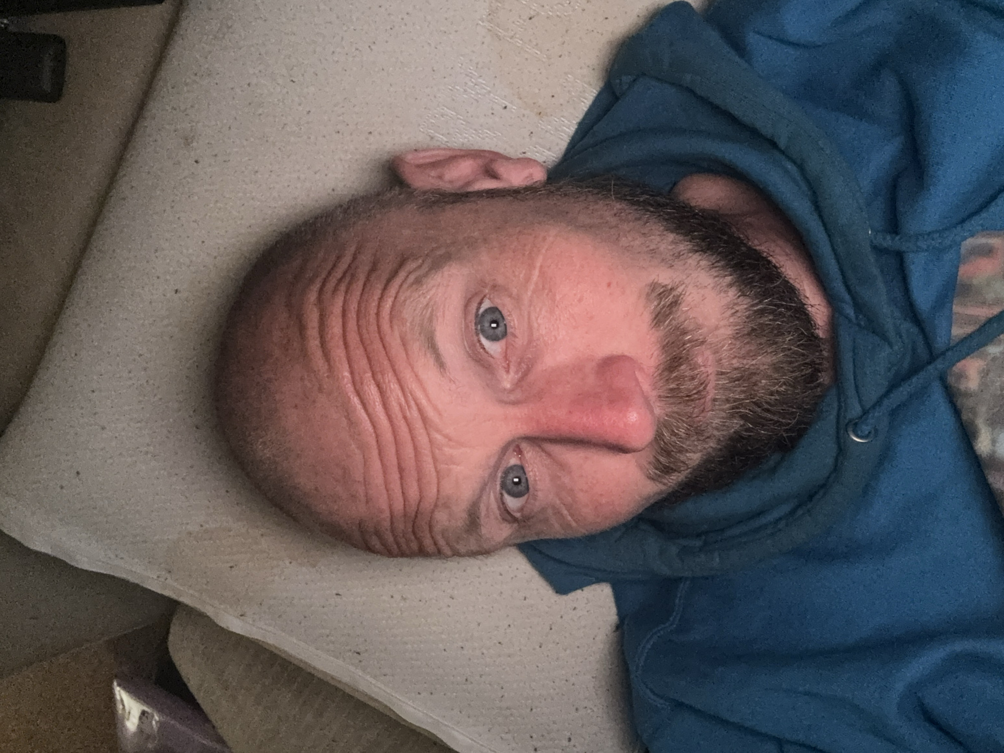 The image is a close-up, overhead selfie of a bald, middle-aged man with blue eyes and a beard, wearing a blue hoodie. He is lying on a textured, off-white surface that appears to be a mattress or cushion. The man's head is tilted, and his facial expression is neutral. The lighting is dim, creating shadows on his face and highlighting the wrinkles on his forehead. The background consists of the same off-white surface and a darker area that could be furniture or a wall. There is no discernible indication of the city of Cheltenham, United Kingdom, or any specific location within it.