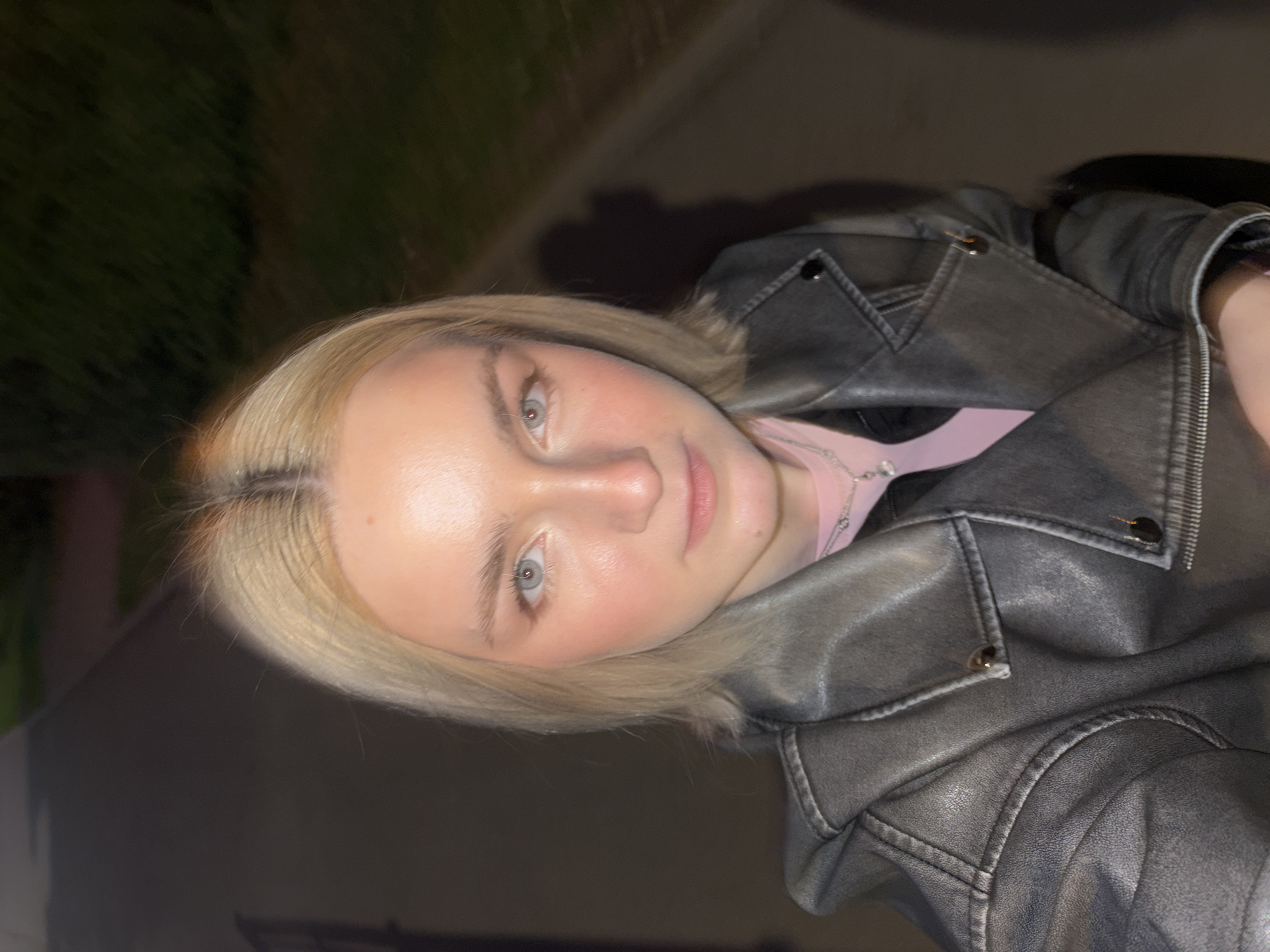 An eye-level, medium shot captures a young Caucasian woman with shoulder-length blonde hair and light blue eyes, facing the camera directly. She is wearing a gray faux leather jacket over a pink shirt and a delicate silver necklace. The woman's makeup includes blush and eyeshadow, and her skin has a slight sheen.
The background is a dimly lit outdoor setting, suggesting it is nighttime. A paved path with a slight incline is visible to the right of the woman, bordered by dark foliage. To the left, a darker area may indicate a building or a shadowed area. The lighting appears to be artificial, casting a soft glow on the woman's face and creating a subtle shadow of her head and shoulders on the path behind her. There are no other people or significant objects in the immediate vicinity. The scene evokes a feeling of urban nightlife, possibly taken while walking or standing on a street or park path. The presence of the paved path and manicured foliage suggests an urban environment like the city of Saint Petersburg, Russia, which is known for its well-maintained public spaces and abundant greenery.