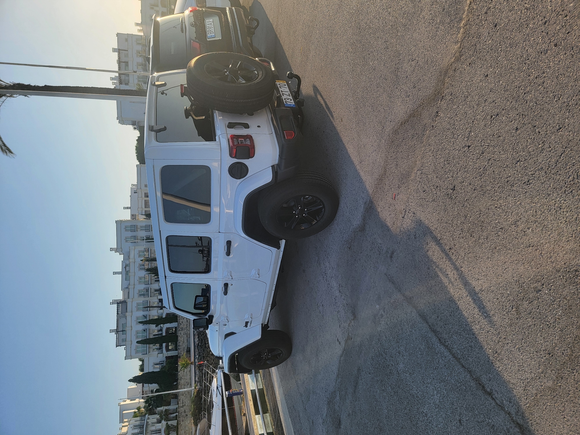 A white four-door Jeep Wrangler is parked on an asphalt surface. The vehicle is oriented to the right, with its rear and right side visible. The Jeep has black rims and a spare tire mounted on the rear. Next to the Jeep, to its right, is a darker colored SUV. In the background, there are white buildings, palm trees, and a clear blue sky. The scene appears to be in Quarteira, Portugal, possibly near a marina, as a boat is partially visible on the left side of the image. The lighting suggests it is either early morning or late afternoon.