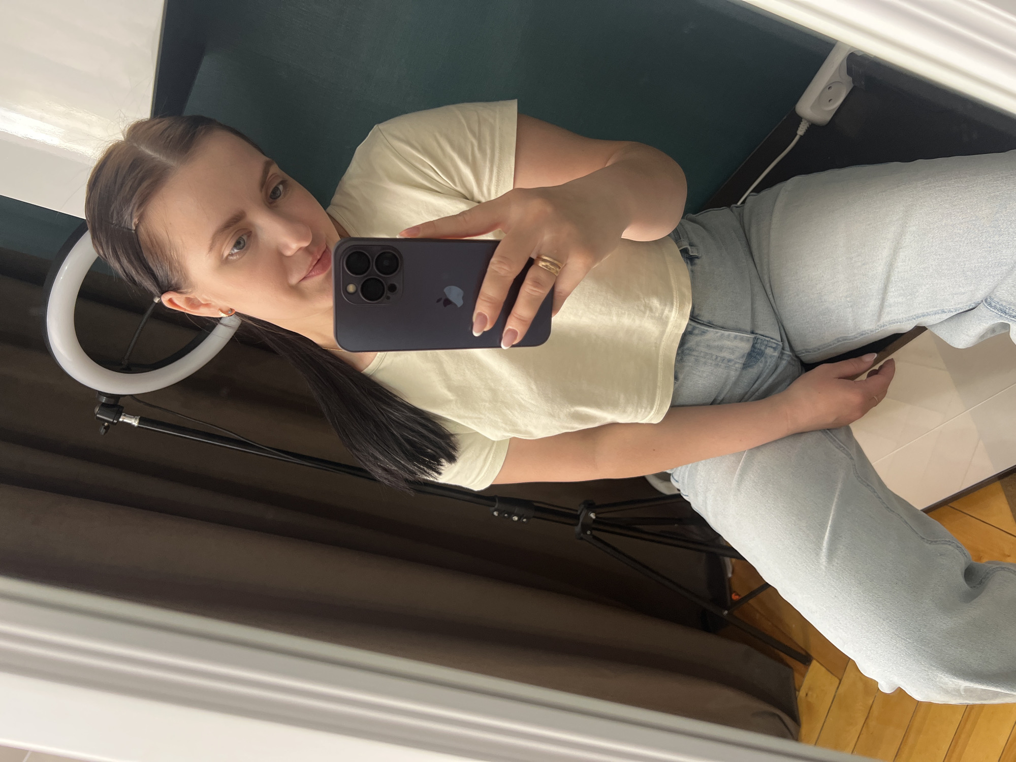 A woman is taking a selfie in what appears to be a room with a dark green wall, white trim, a ring light, and a light-colored floor. She is holding a dark-colored iPhone in her right hand, with a gold-colored ring visible on her finger. She is wearing a cream-colored t-shirt and light blue jeans. The location may be Марковский, Russia, but there is no definitive evidence to confirm this.