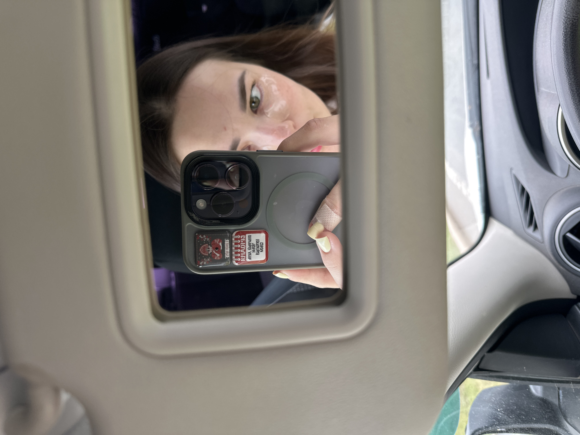 A young woman with brown hair is seen in a car's vanity mirror, taking a selfie with her phone. She has under-eye patches on and is wearing a light-colored shirt. Her phone has a gray case with two stickers on it. One sticker features a red mushroom character, and the other is a white label with red text. She has light yellow nail polish on her fingernails and a bandage on her left index finger. The car interior is visible, with the dashboard and steering wheel in the foreground. The exterior view through the car window appears to be a grassy area. There is no specific information about the city of Perm, Russia in the image or its metadata.