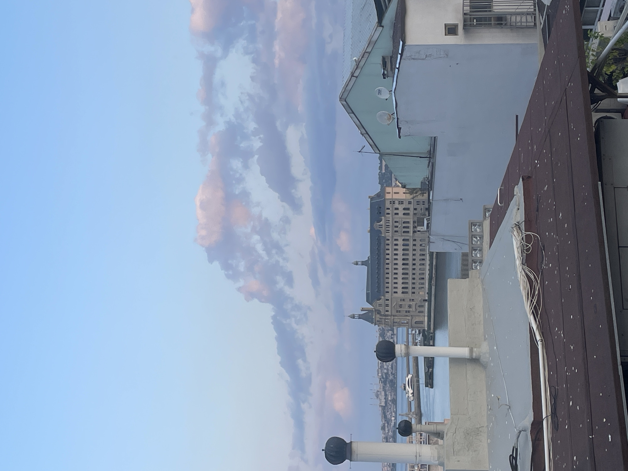 This photograph captures a view of the Kadıköy district of Istanbul, Turkey, from a rooftop. The foreground is dominated by the roofs of residential buildings, with a dark brown, sloped roof in the lower section featuring scattered white debris. Above this, a lighter gray, flat roof is visible, partially obscured by wires and pipes.
In the middle ground, two prominent white structures with black caps rise from the rooftops, likely chimneys or ventilation shafts. Beyond these, the blue waters of the Bosphorus strait are visible, with a white ferry boat traversing the water. Further in the distance, a large, historic building with a mansard roof and numerous windows stands prominently. This is likely the Haydarpaşa Terminal. The skyline of Istanbul, including the silhouette of mosques with minarets, can be seen in the far distance.
The sky above is a soft blue, with scattered clouds tinged with pink and purple, suggesting either dawn or dusk. The overall atmosphere is calm and serene. The image is devoid of any visible people or direct human interaction. The scene is an architectural and urban landscape, focusing on the built environment and its relationship with the natural elements of water and sky.