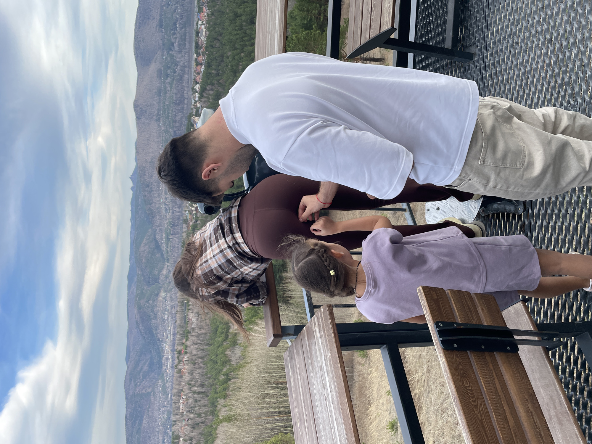 Captured from an elevated vantage point in Krasnoyarsk, Russia, the image presents a family of three—a man, a woman, and a girl—all oriented towards the mountainous and cityscape panorama sprawling in the background.
The man, donned in a white t-shirt and khaki pants, leans forward, his gaze directed downwards, presumably observing something below. The woman, wearing a brown and plaid shirt, also leans forward towards the edge. The girl, clad in a lavender dress, is also leaning towards the edge to see what is below.
They are on a platform or balcony constructed with metal grating for the floor and railing with wooden benches. In the background, the landscape reveals a mix of mountainous terrain and urban development. A blend of evergreen forests and residential structures are seen. The sky above is partly cloudy.