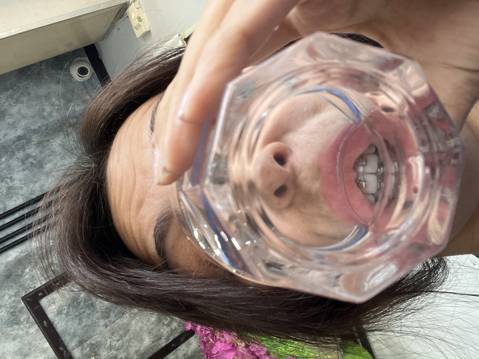A person is holding a clear, multifaceted glass up to their face, with their nose and mouth visible through the glass. The person has brown hair. The background includes a gray tiled floor, a white cabinet, and what appears to be a surveillance camera mounted on the ceiling. The setting is likely indoors, but a precise location in Kuta Selatan, Indonesia, cannot be determined from this image.