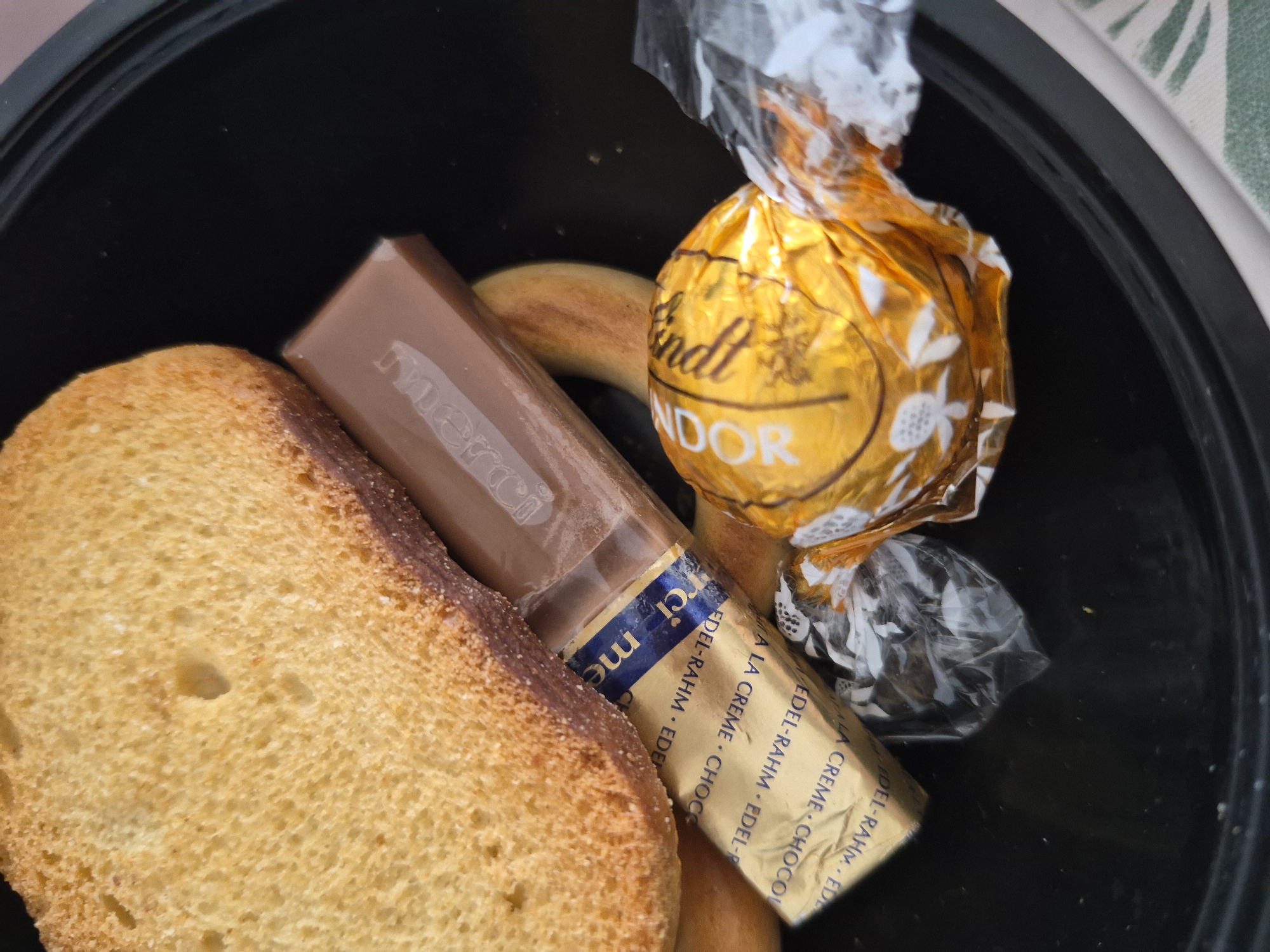 A close-up shot shows a black bowl containing several food items. In the upper left corner, a slice of toasted bread with a golden-brown crust and a porous interior is visible. To the right of the bread, a dark brown chocolate bar with the brand name "Merd" embossed on it lies diagonally. Below the chocolate bar, a small, rectangular gold foil-wrapped item with blue text, possibly a butter or cream packet, is positioned. Partially obscuring the foil packet is a round, gold-foil-wrapped Lindt chocolate with white floral patterns. The wrapper is crumpled, and some of the silver plastic wrapping of another item is visible to the right. The background is a plain black surface, consistent with the interior of the bowl. The image provides no information about its location in the city of Unknown, Unknown, nor does it depict any people or actions.