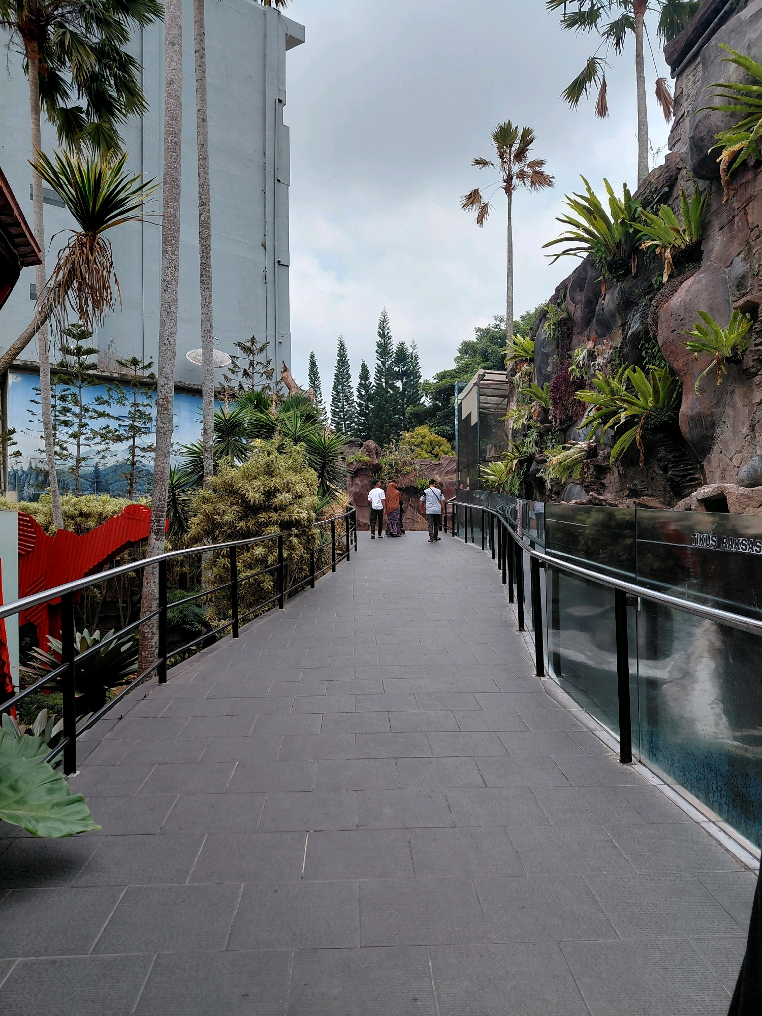 The image depicts a walkway with dark gray square paving stones, lined by a metal railing with black posts and a glass barrier. To the left of the walkway is a tall, light blue building with some palm trees and other greenery in front of it. A large red structure is partially visible on the far left. To the right of the walkway, a rock-like wall is adorned with lush green ferns and other plants. The background shows a cloudy sky and some tall evergreen trees. Three individuals are walking away from the camera on the walkway. The sign "TIKUS RAKSASA" is visible on the glass barrier to the right. This scene appears to be within the Jatim Park 2 zoo in Batu, Indonesia, known for its "Giant Rat" exhibit.