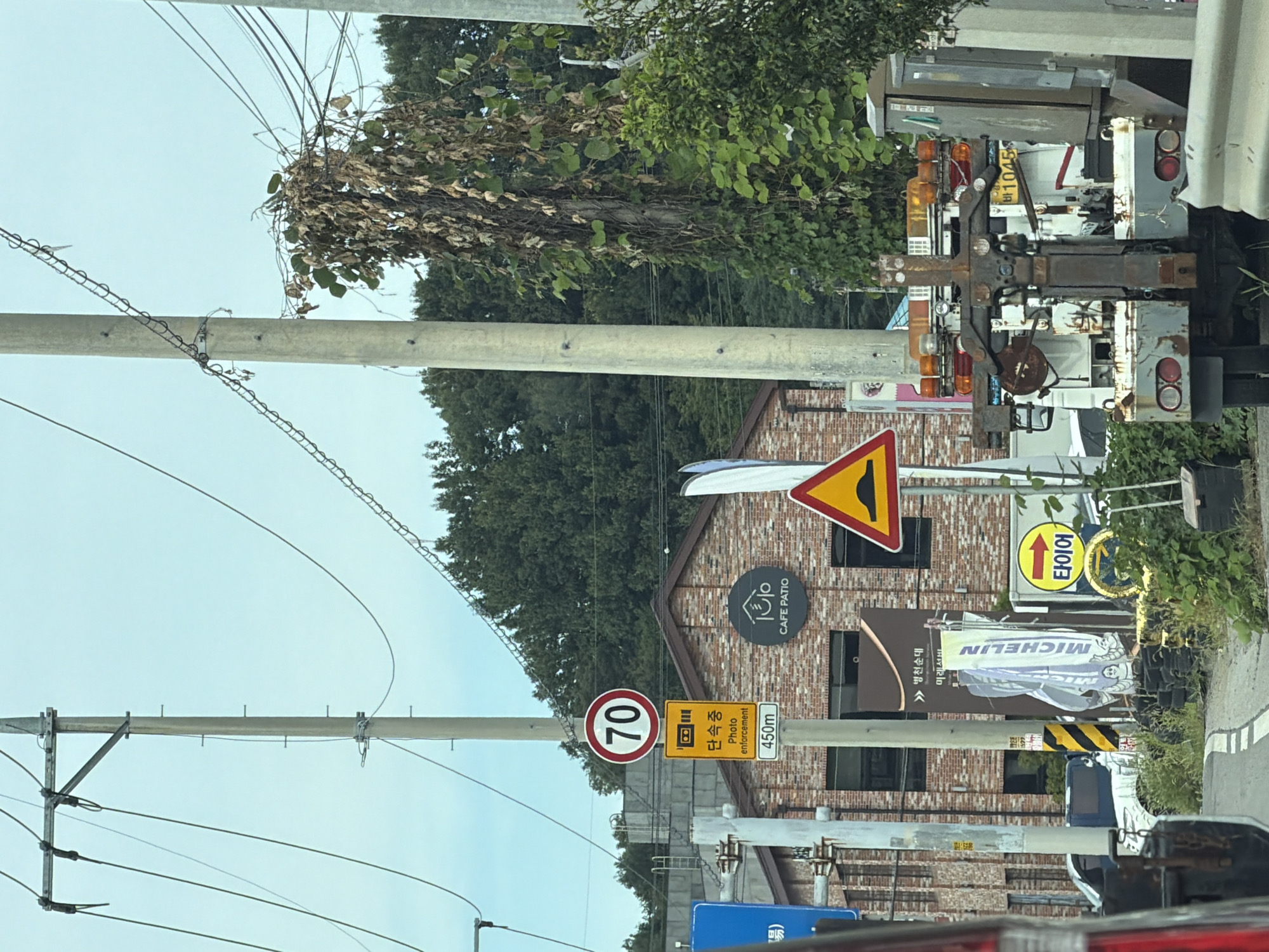 The image captures a roadside scene in Hwaseong-si, South Korea, under an overcast sky. A concrete utility pole stands prominently, supporting electrical wires and adorned with traffic signs. One sign indicates a speed limit of 70, another warns of photo enforcement at 450m, and a third, triangular sign depicts a speed bump. A brick building with "CAFE PATIO" visible on its facade is situated behind the pole. To the right, a tow truck is parked, its equipment extended. Further down the road, a Michelin banner and a yellow sign with an arrow pointing right are visible. The environment is moderately vegetated with trees and bushes lining the roadside. The image appears to have been taken from inside a vehicle, as indicated by the partial view of the vehicle's interior on the left side.