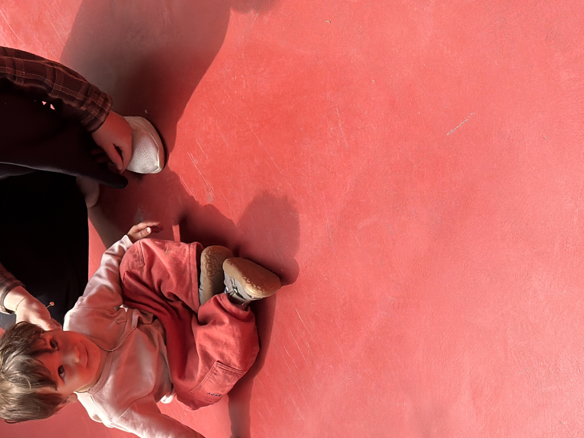 The image shows a child lying on a red surface. The child is wearing red pants and a light-colored shirt. An adult wearing a brown plaid shirt and black pants is partially visible, holding the child's hand. The scene appears to be outdoors, based on the lighting. The location is Yerevan, Armenia.