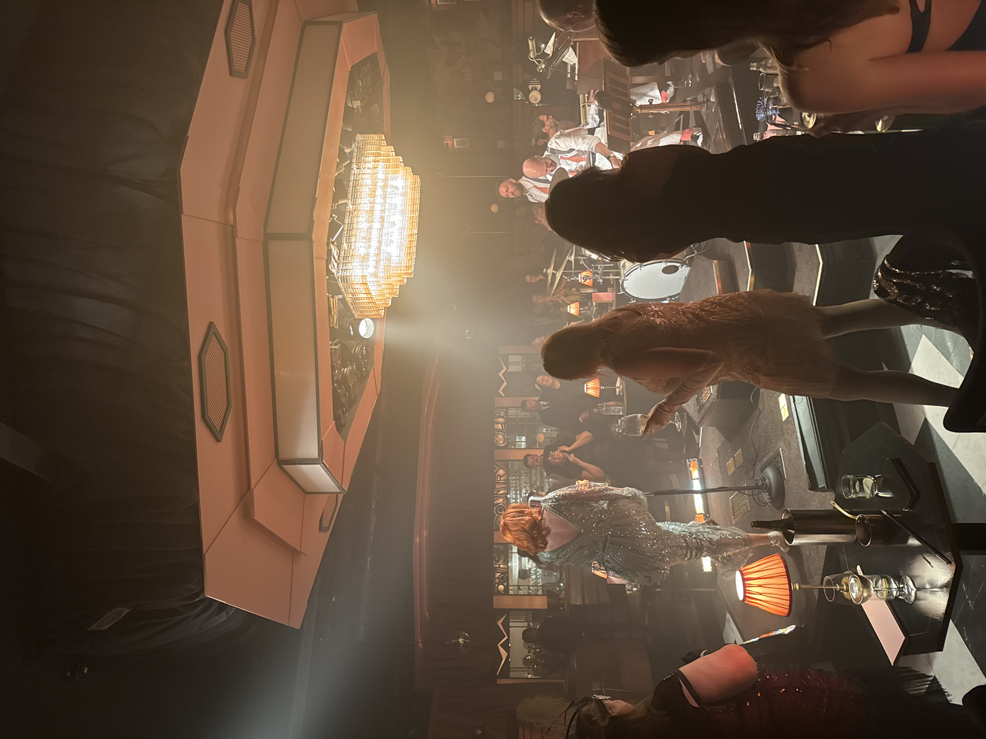 The image depicts a dimly lit interior, possibly a speakeasy or jazz club, with a large, art deco chandelier dominating the left side. Several people are visible, seemingly patrons and performers. A woman in a shimmering silver dress stands near the lower center, potentially a singer or dancer. Behind her, another individual in a gold dress is also visible. Several small tables with drinks and lamps are arranged around the room, indicating a social setting. In the background, a band can be seen performing on a stage. The overall atmosphere suggests an evening of entertainment in a stylish, vintage-themed establishment.