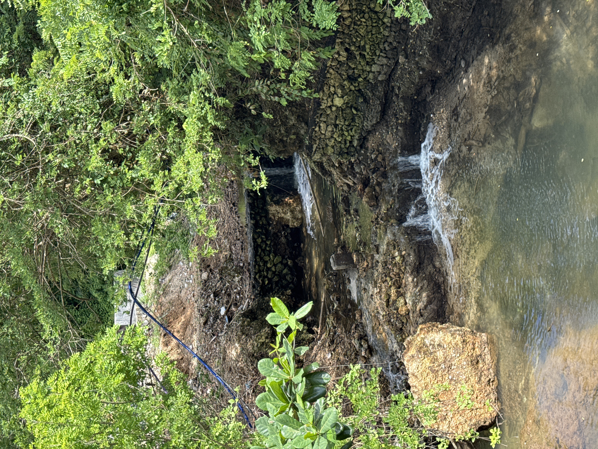 The image shows a small waterfall in Kuta Selatan, Indonesia. The water flows over rocks and concrete structures into a pool. Lush green vegetation surrounds the waterfall, growing on the rocky banks. There is a blue pipe running along the left bank. A large rock sits in the pool at the base of the falls.