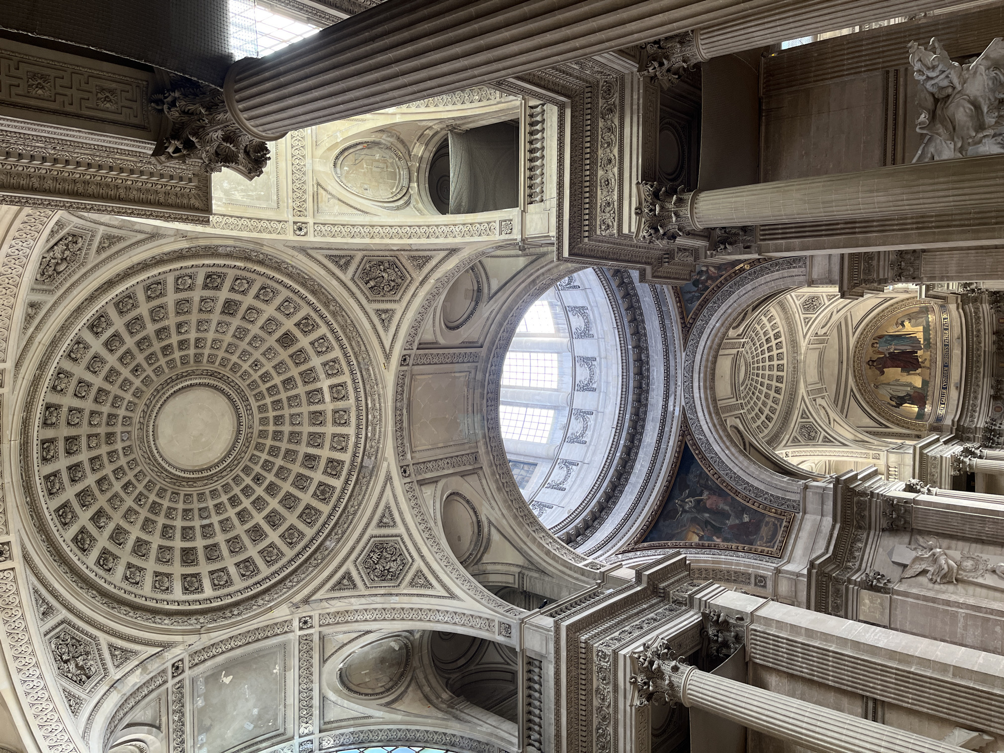 This image captures the grand, ornate interior of a large classical-style building, viewed from a low angle looking upwards. The scene is dominated by elaborate architectural features, including multiple domes, arches, and colossal columns.
A prominent central dome features a detailed coffered ceiling with a radiating circular pattern. Surrounding this are vaulted ceilings, intricate moldings, and relief sculptures showcasing complex decorative motifs and classical designs. Tall, fluted Corinthian columns with richly carved capitals support the upper structures.
Natural light floods the space from large, multi-paneled windows positioned high up in the building, illuminating the detailed craftsmanship and creating a sense of height and grandeur. The lighting suggests it is daytime.
Murals and paintings adorn some of the curved surfaces and archways. On the right side, a notable mural depicts several figures, possibly religious or historical, rendered in warm tones with visible gold accents. Above this mural, Latin text is discernible: "CONSOLAVIT CONSOLATRIX PAUPERUM" (The comforter of the poor comforted) and "SIT NOMEN DOMINI" (Blessed be the name of the Lord).
The color palette is predominantly cool, consisting of various shades of stone, grey, cream, and off-white, emphasizing the classical marble and stucco work. No people are visible, allowing the focus to remain entirely on the architectural and artistic elements. The overall impression is one of immense scale, intricate detail, and historical significance. The architecture and Latin inscriptions are characteristic of the Pantheon in Paris, France.