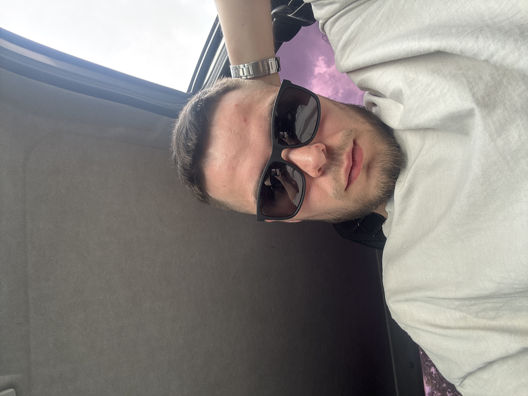 The media file is a close-up, slightly angled, portrait-style photo of a young man wearing sunglasses. The man is looking towards the camera, with a neutral expression. He has short, dark hair and a beard. He is wearing a light-colored t-shirt and a silver watch on his left wrist. The background is blurred and appears to be the interior of a vehicle, possibly a car, with a sunroof open, revealing a sky with purple and pink hues, suggesting either dawn or dusk. The overall lighting suggests it is daytime. The image's orientation is rotated, with the top of the frame appearing to the left. No visible text is present in the image.