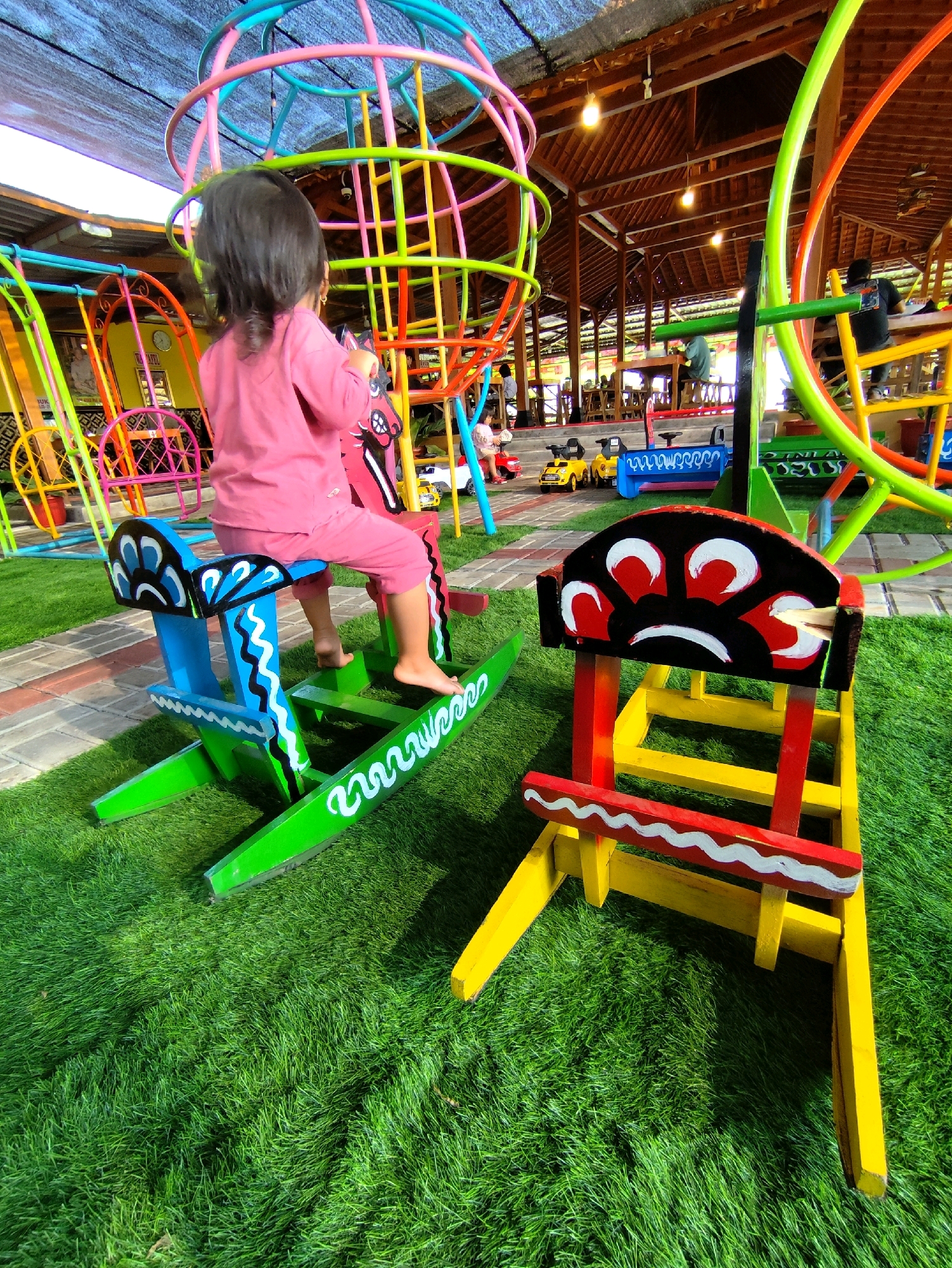 A young child, wearing a pink long-sleeved shirt and pink pants, sits on a brightly painted wooden rocking horse. The horse is predominantly red and blue with white and black decorative accents. The child's feet are bare and rest on the green rocking mechanism. In the foreground, another wooden seat, painted red and yellow with white wavy patterns and black and red circular designs, is visible. The ground is covered in vibrant green artificial turf. Behind the child and the rocking horse, a large, colorful metal jungle gym structure with rings and bars in various pastel colors dominates the background. The area appears to be an outdoor playground or recreational space, possibly part of a restaurant or resort, given the covered seating and wooden beams of a building visible in the upper portion of the image. The overall impression is a lively and colorful children's play area. This scene is located in Klaten, Indonesia.