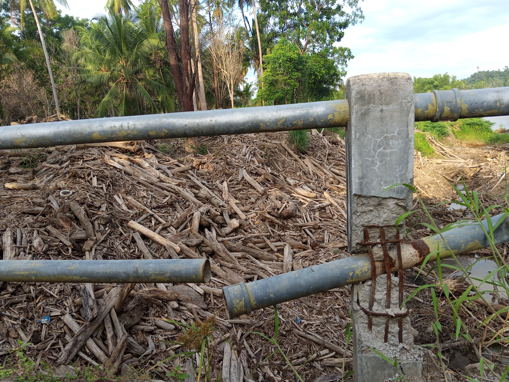 The scene in Kruengbeukah, Indonesia, depicts a landscape cluttered with driftwood and logs. A dilapidated metal pipe fence runs across the foreground, supported by crumbling concrete posts with exposed rebar. The lower pipe is broken. Behind the fence is a large pile of wood debris. The background consists of various trees, including palm trees, and some greenery near a body of water. The sky is partially cloudy.