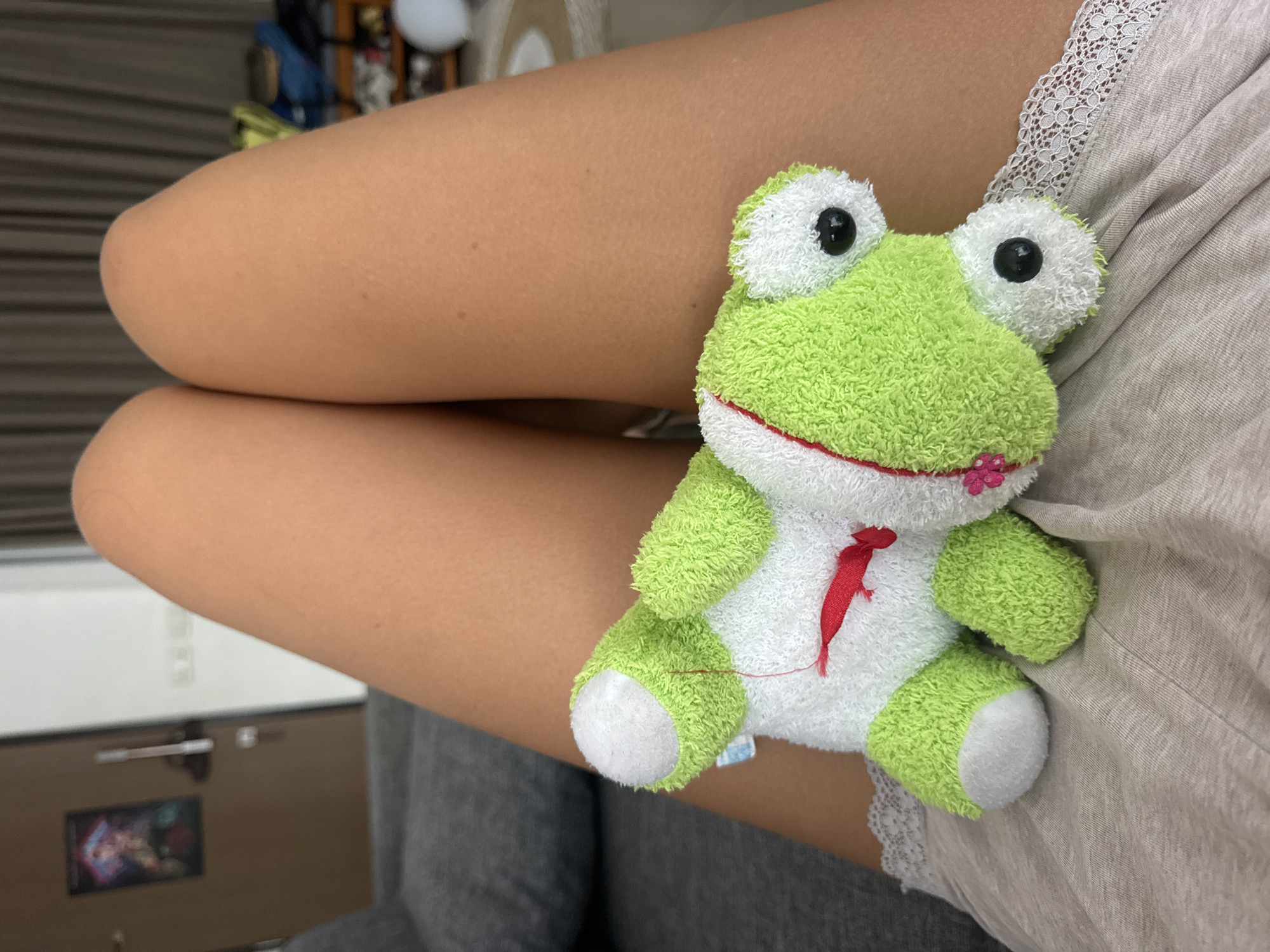 A stuffed frog toy sits on a person's lap. The frog is green and white with black button eyes and a red embroidered tie. The person's bare legs are crossed, and they are wearing a light gray garment with lace trim. The setting appears to be indoors.